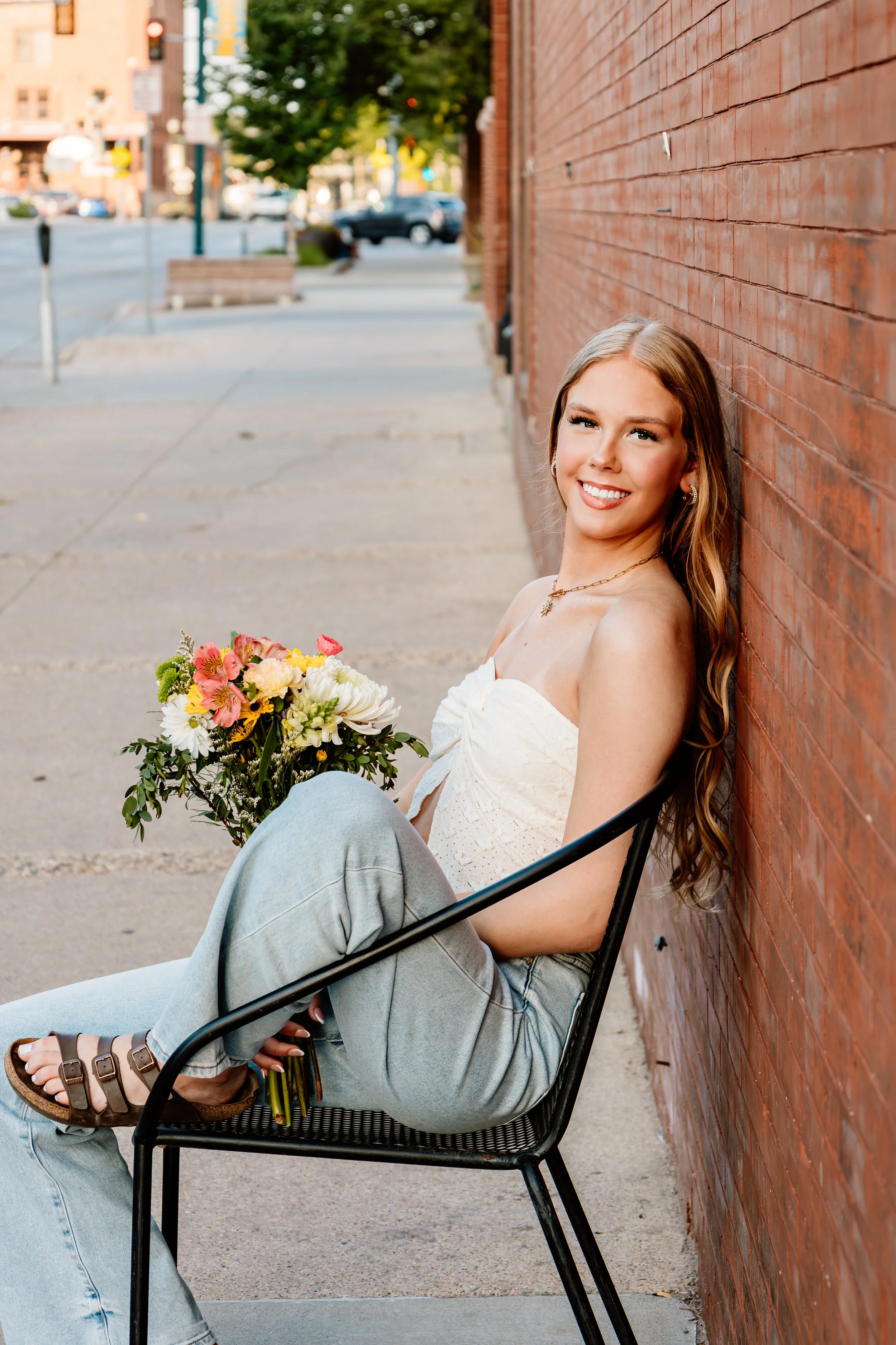 A young woman with long wavy hair sitting on a black chair against a brick wall on a city street, holding a bouquet of flowers, smiling at the camera in Downtown Sioux Falls, South Dakota.