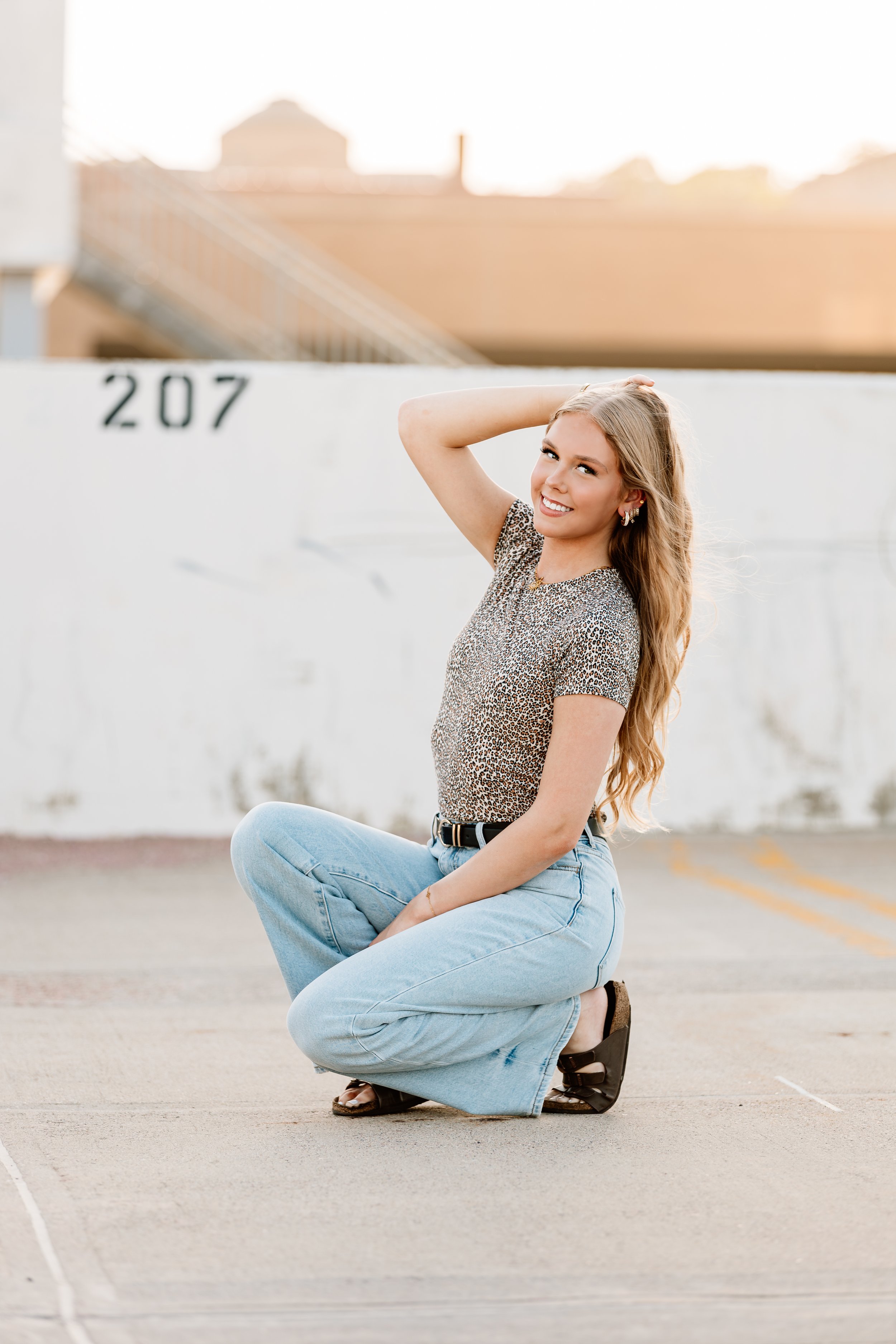 A young woman with long blonde hair, wearing a leopard print top, light blue jeans, and black sandals, kneeling on concrete ground in an outdoor parking lot at sunset, smiling at the camera in Downtown Sioux Falls, South Dakota.