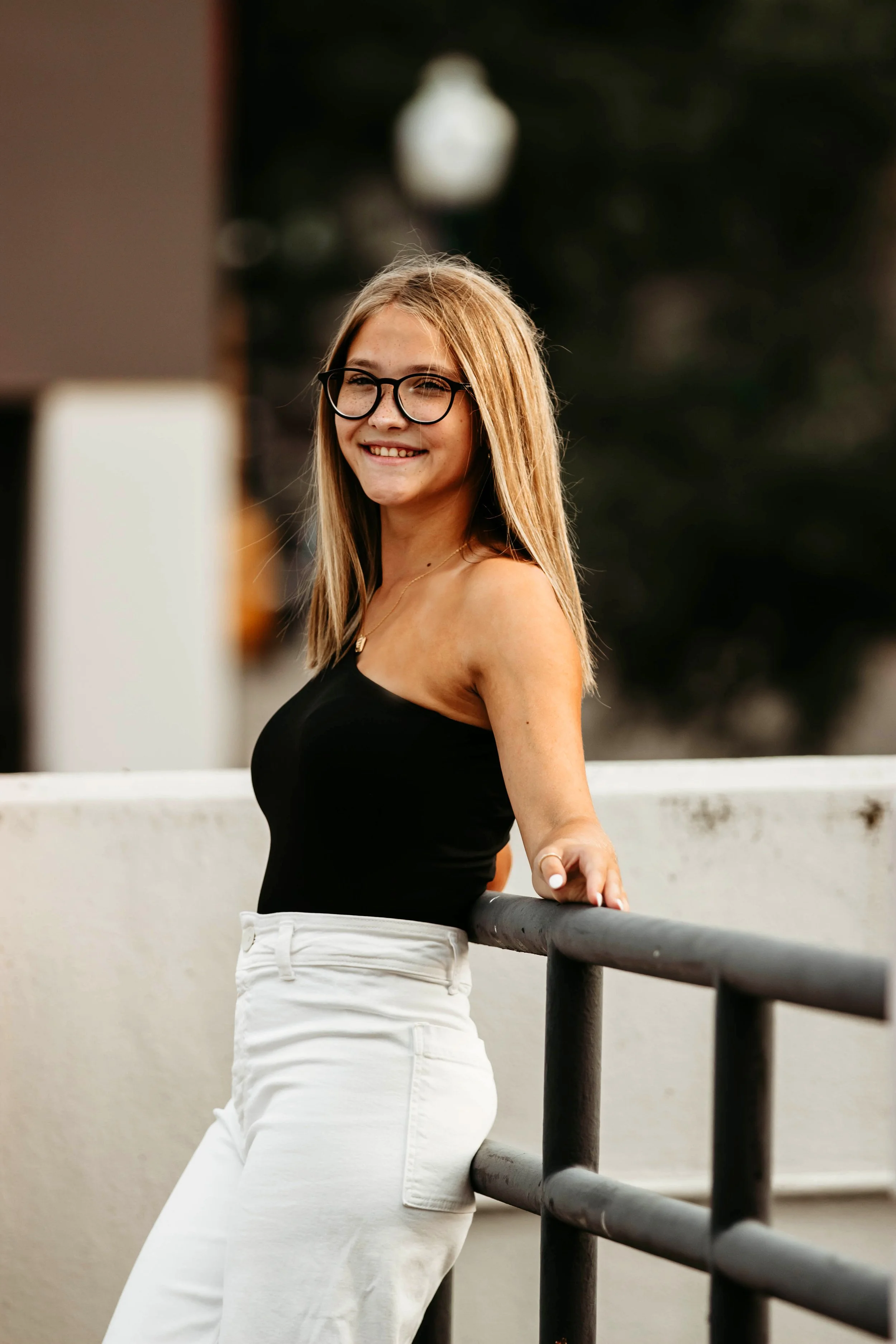 A young woman with blonde hair, glasses, and a black strapless top, smiling and leaning against a black railing outdoors in Downtown Sioux Falls, South Dakota.