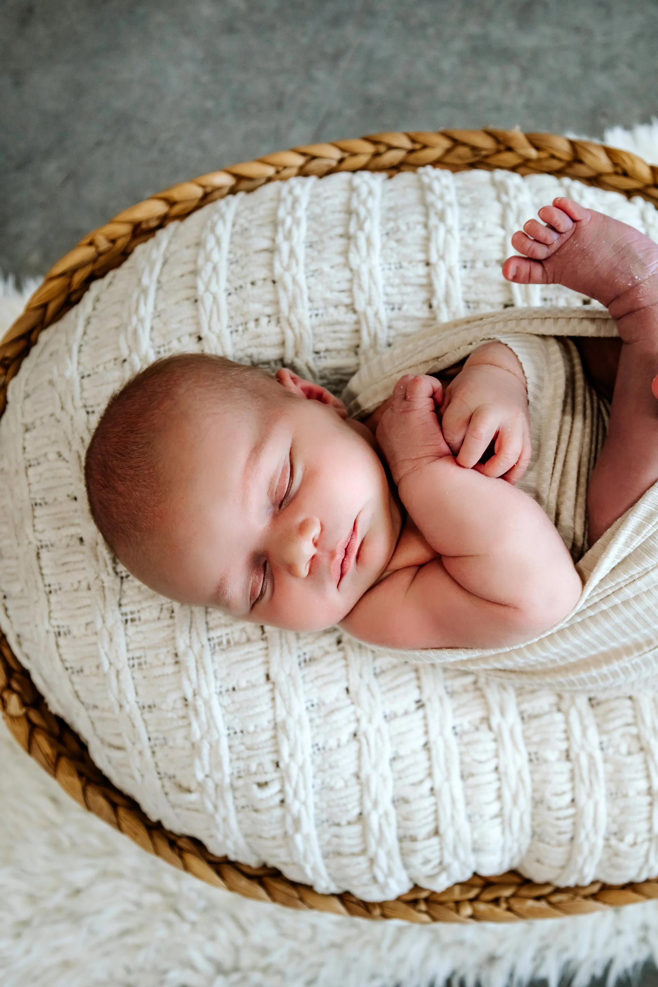A sleeping baby lying on a white knitted blanket in a wicker bassinet, with a hand gently touching the baby's foot at The White Space Studio in Sioux Falls, South Dakota.