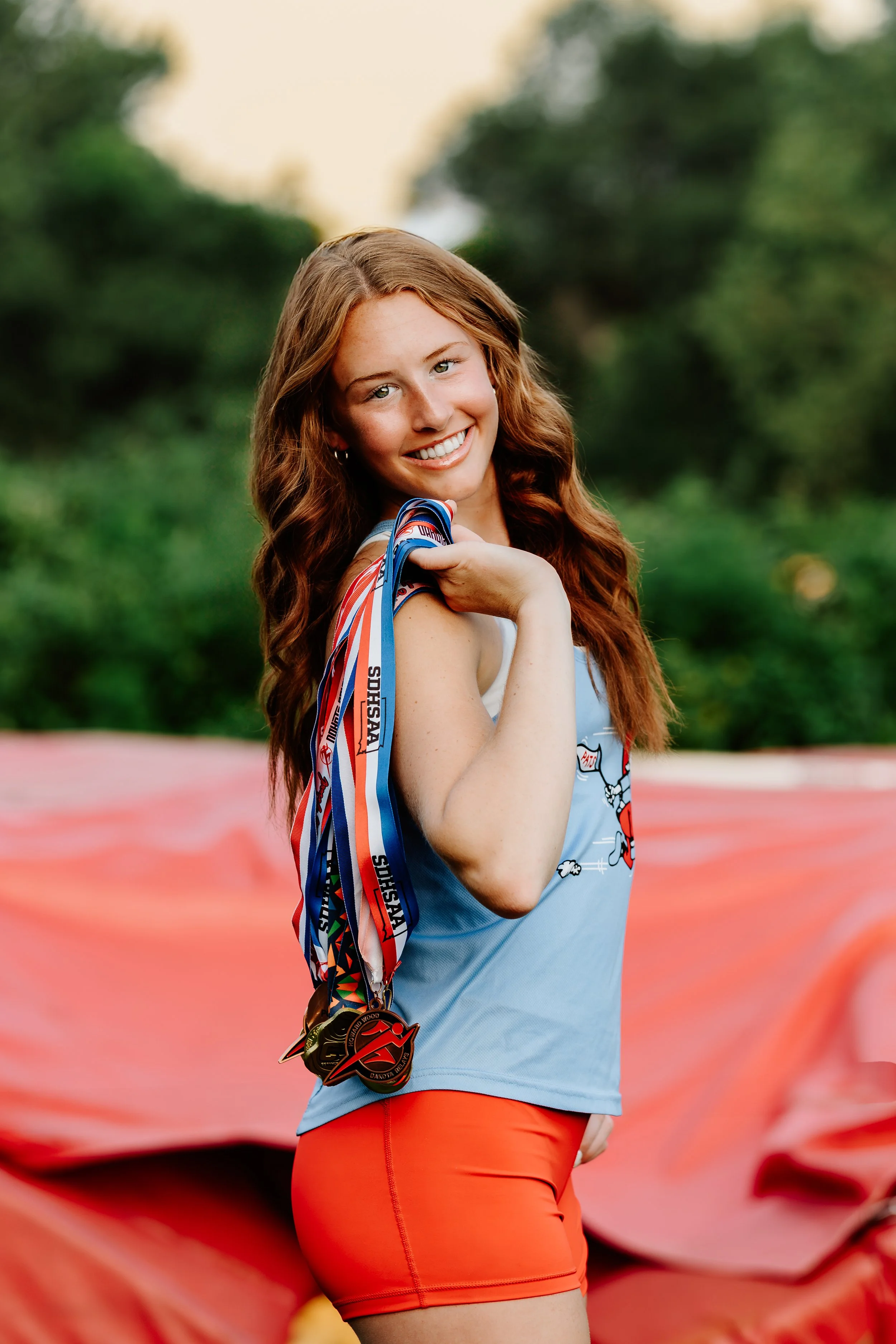 A young woman with long, wavy red hair smiling and holding medals on a ribbon over her shoulder, standing outdoors with green trees and a pink fabric background at Good Earth State Park in Sioux Falls, South Dakota.
