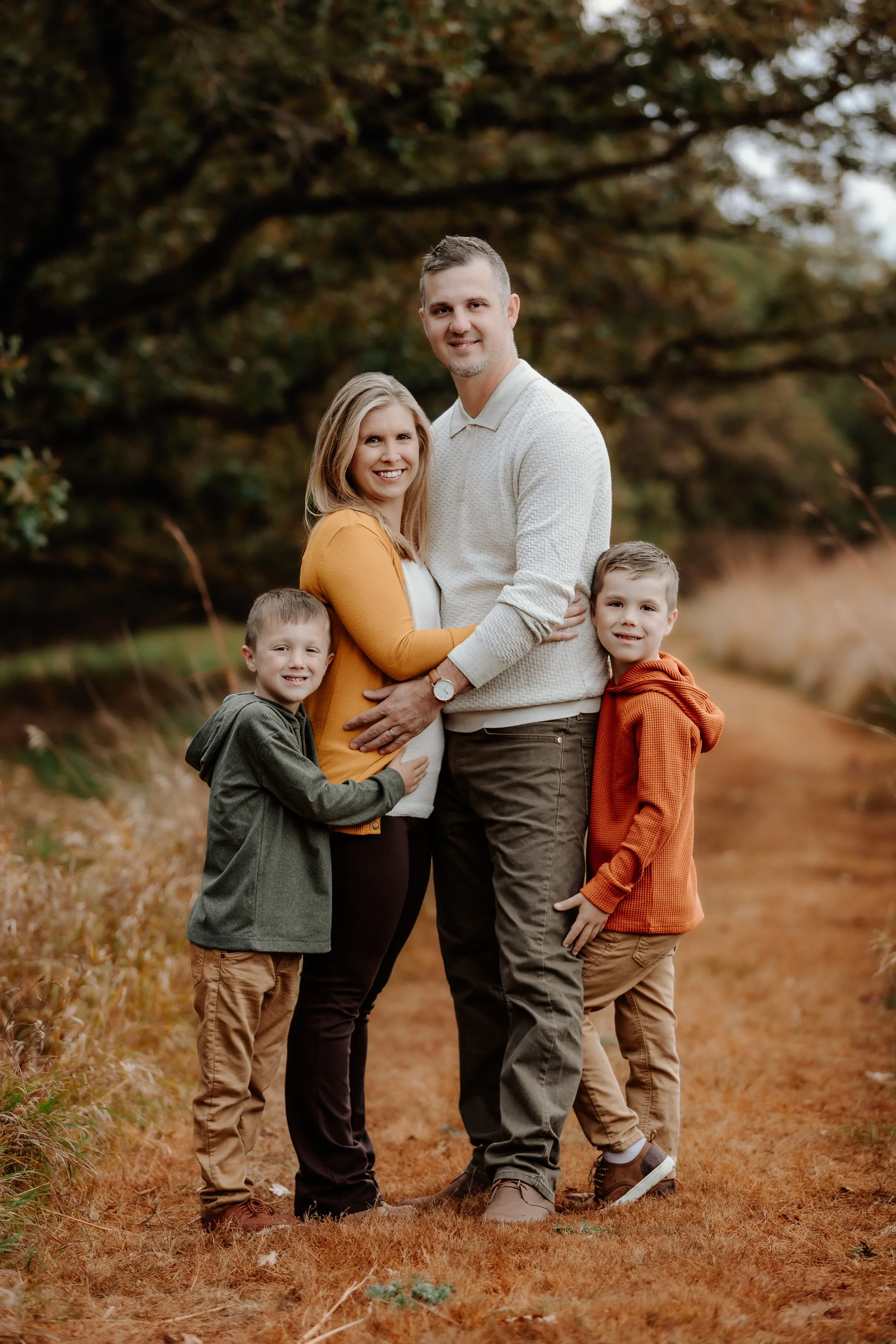 A family of four standing outdoors in autumn, with trees and fallen leaves in the background. The pregnant mother is in the center, wearing a white top and yellow cardigan, smiling in Sioux Falls, South Dakota at Good Earth State Park.