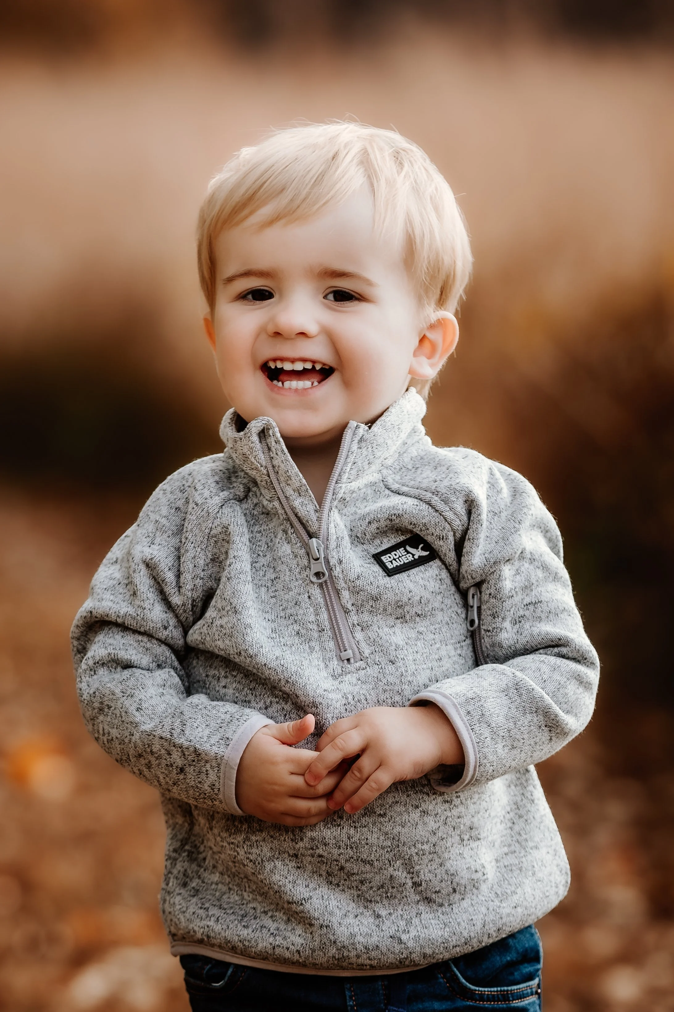 A smiling young boy with blonde hair, wearing a gray sweater, standing outdoors on a fall day with blurred brown background in Sioux Falls, South Dakota at Good Earth State Park.
