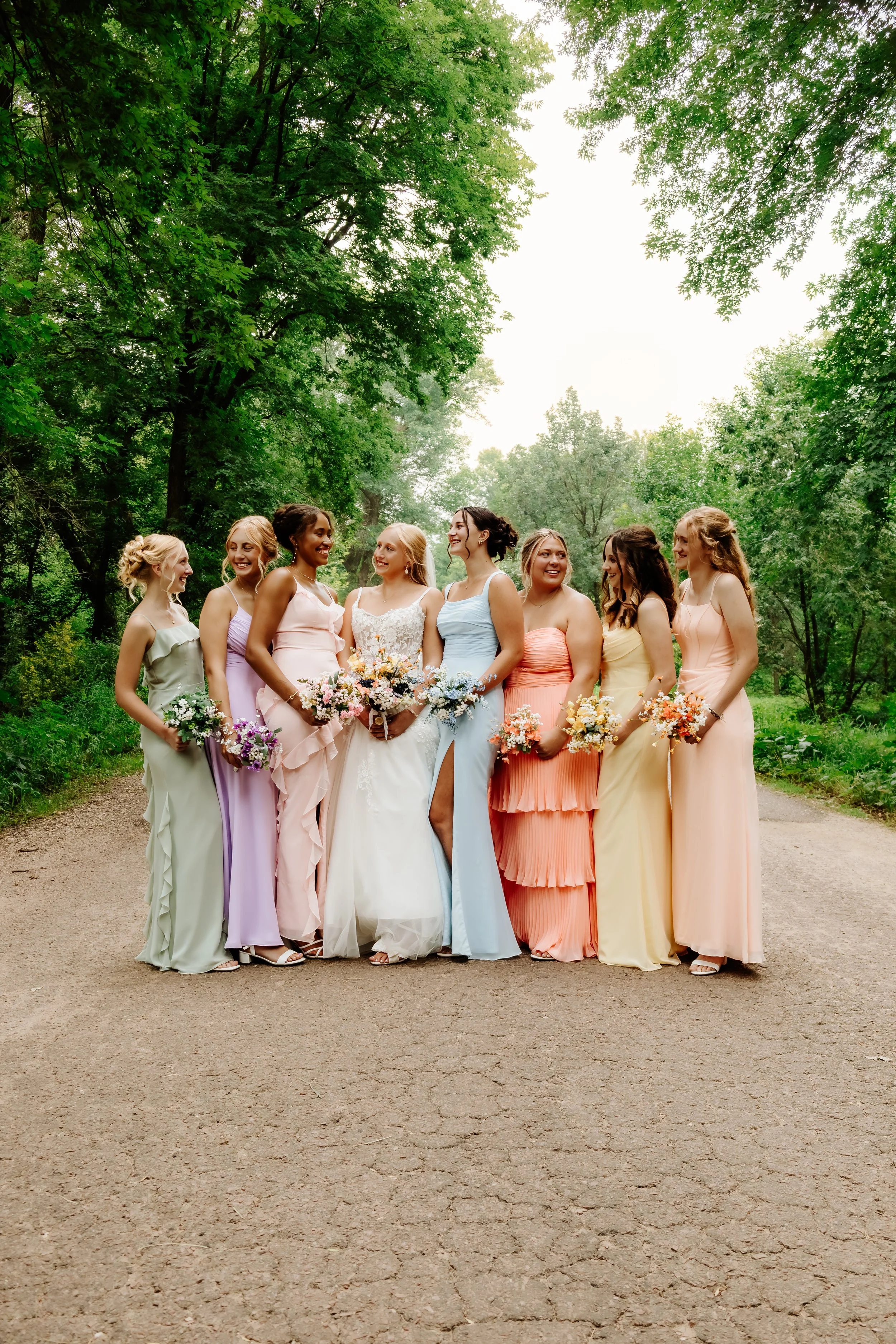 A group of nine women, including a bride in a white wedding gown, standing on a dirt path surrounded by green trees, holding colorful bouquets, during a daytime outdoor wedding photoshoot in Luverne, MN.