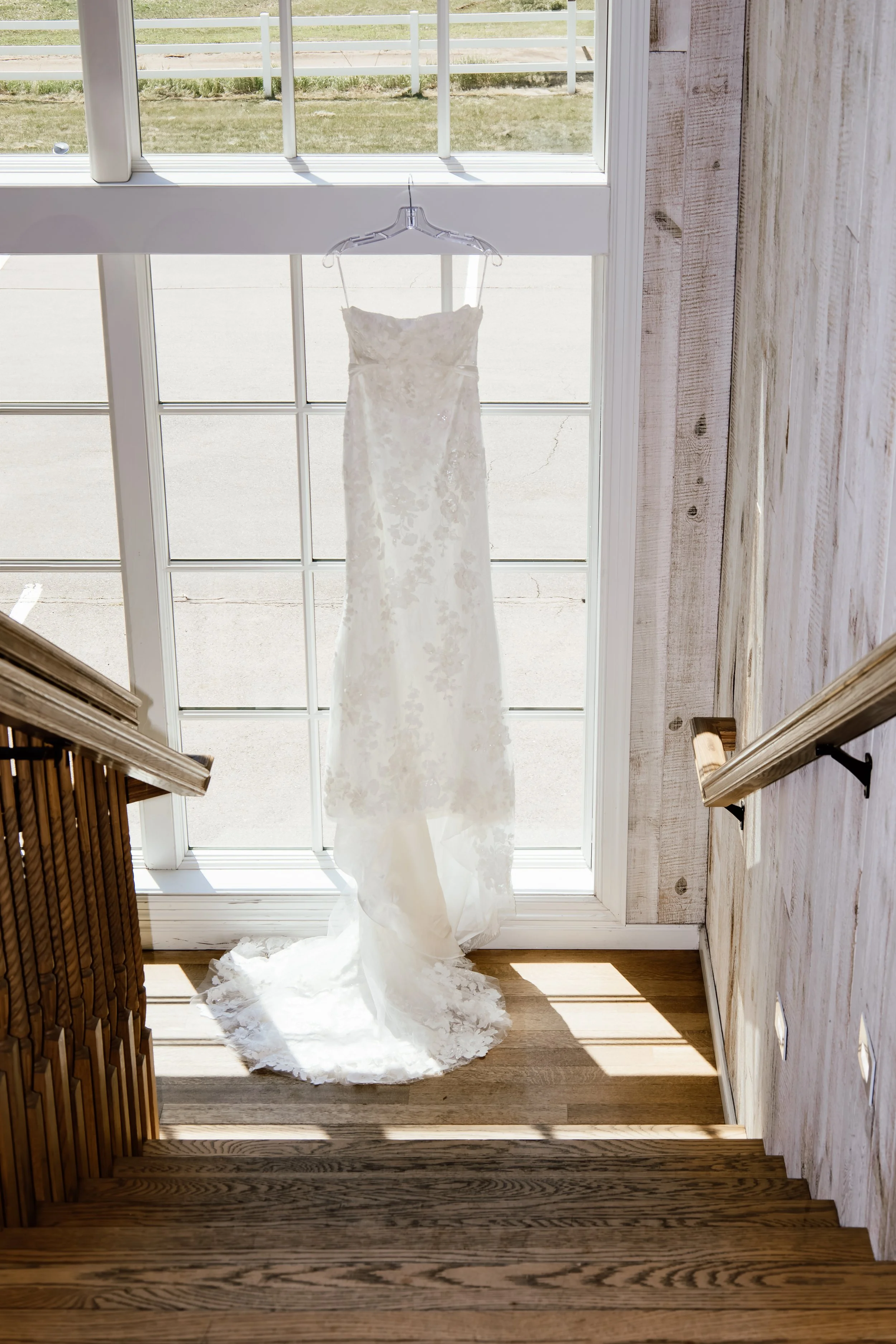 A white wedding dress hanging on a hanger in front of a large window, with sunlight shining through. The dress has lace details and is positioned on a hanger attached to a window frame at Laurel Ridge in Sioux Falls SD.