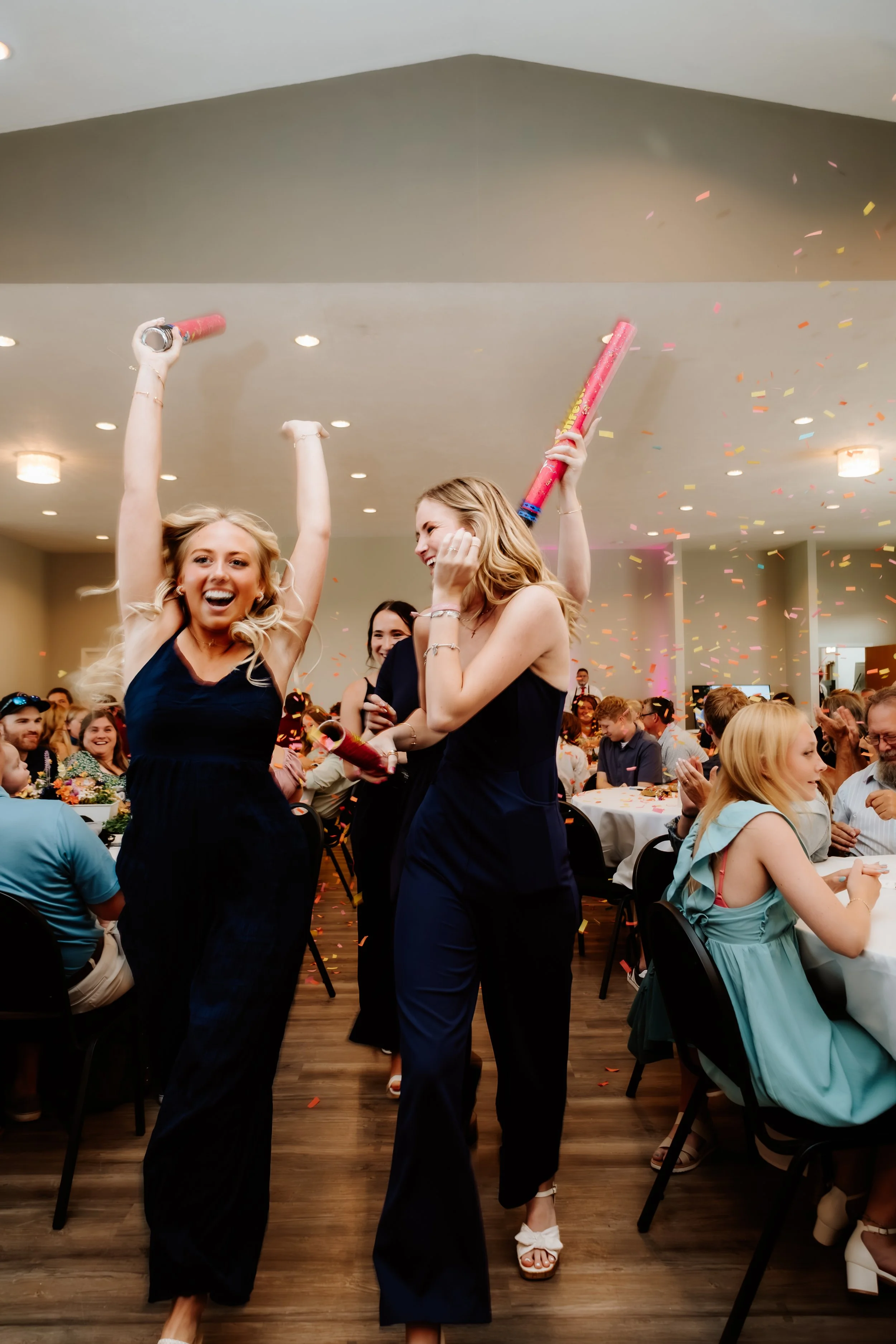 Two women celebrating at a party, holding confetti poppers, with confetti falling around them and guests seated at tables in the background in Luverne, MN.
