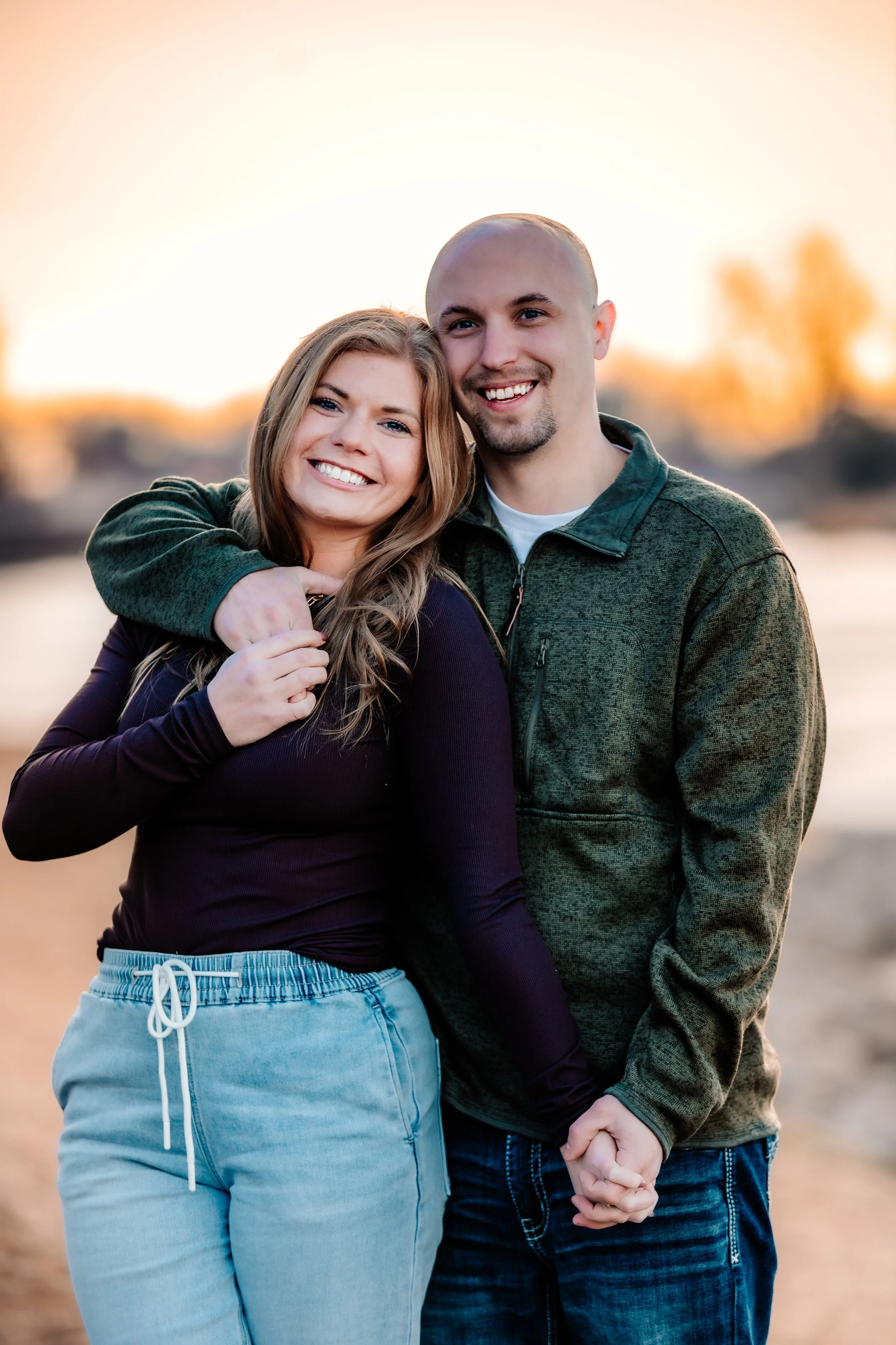 A happy couple holds hands and embraces outdoors during sunset, with a blurred natural background at Terrace Park in Sioux Falls, South Dakota.