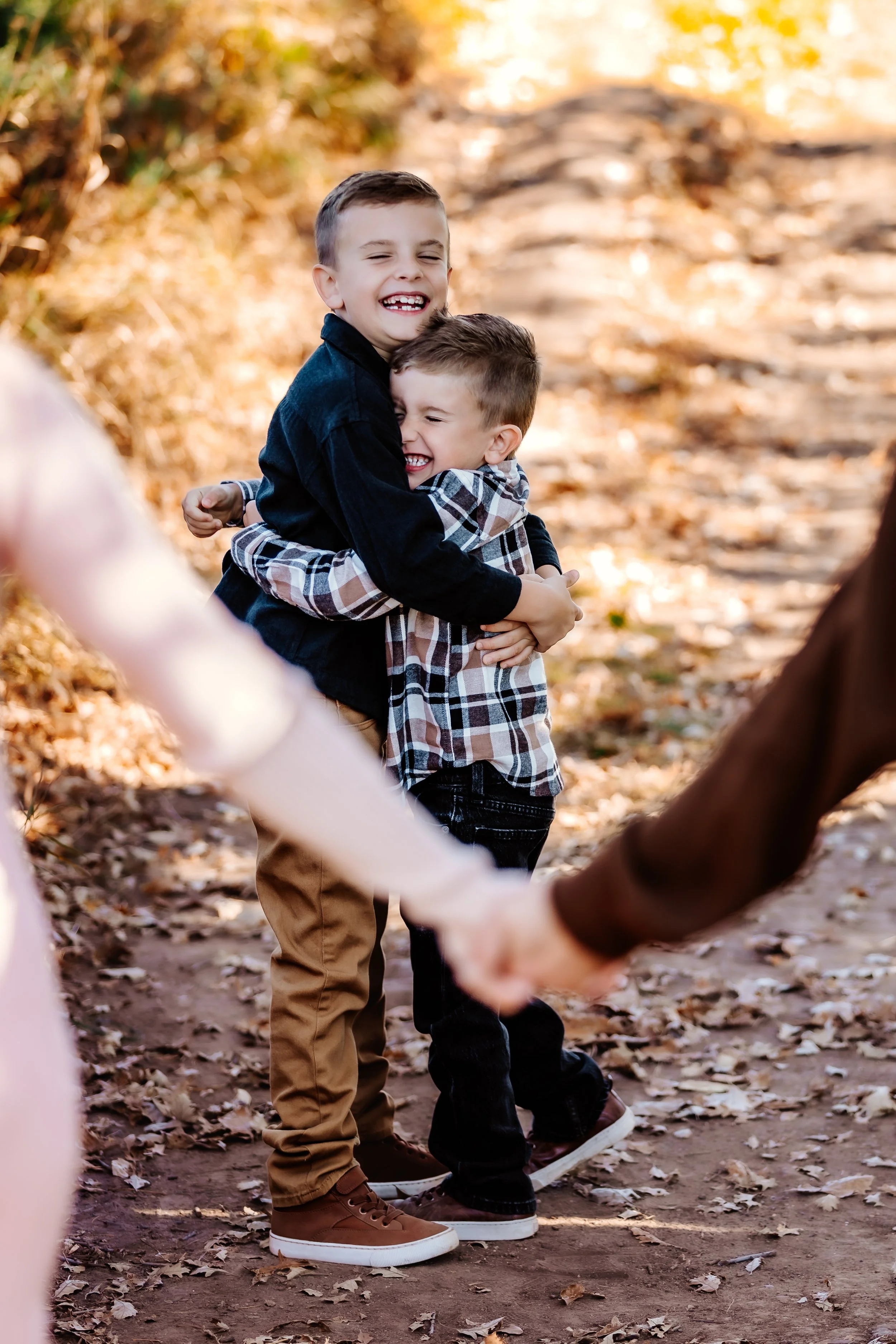 Two young boys hugging each other and smiling while holding hands with another pair of people in a wooded area with fallen leaves in Sioux Falls, South Dakota at Good Earth State Park.