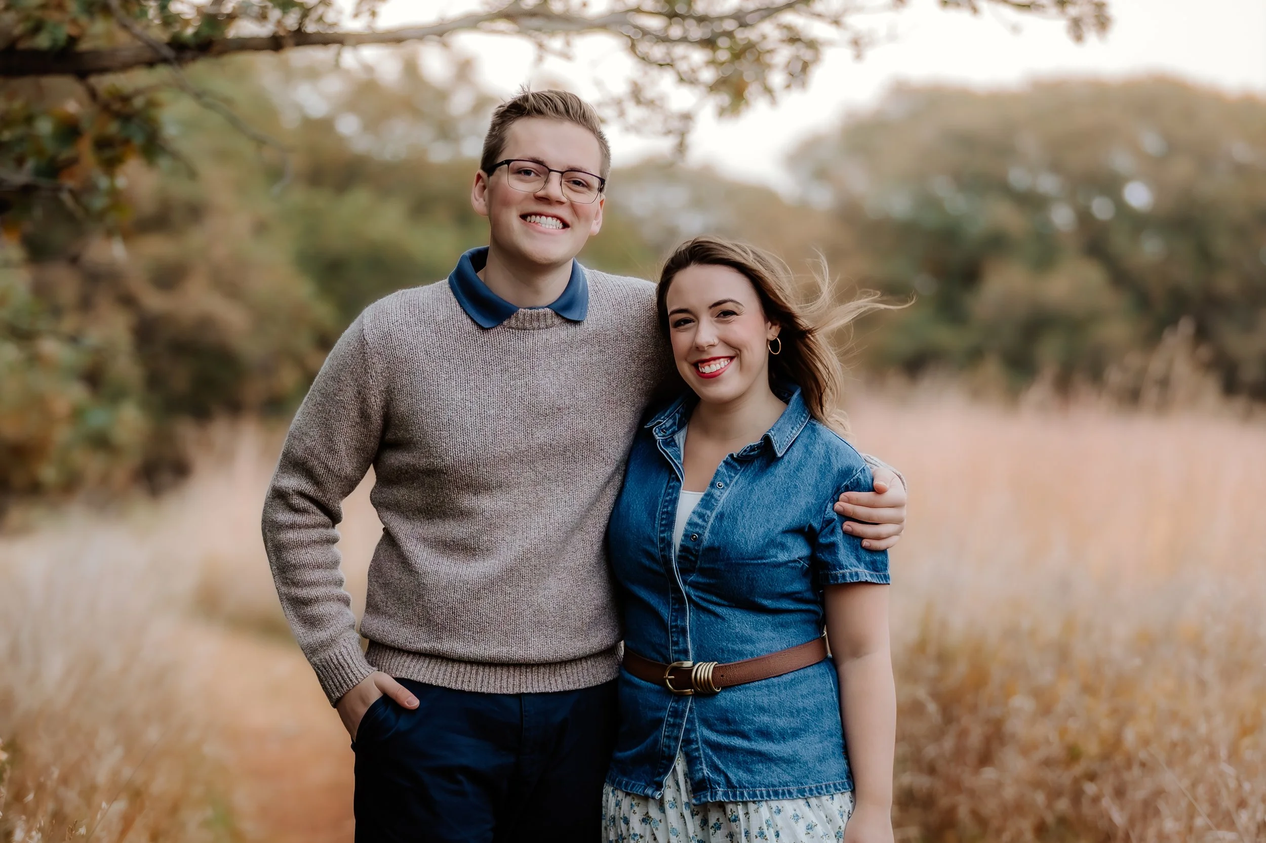 A smiling young man wearing glasses and a beige sweater stands next to a smiling young woman in a denim dress with a brown belt, outdoors in a field with trees and golden grass in the background in Sioux Falls, South Dakota at Good Earth State Park.