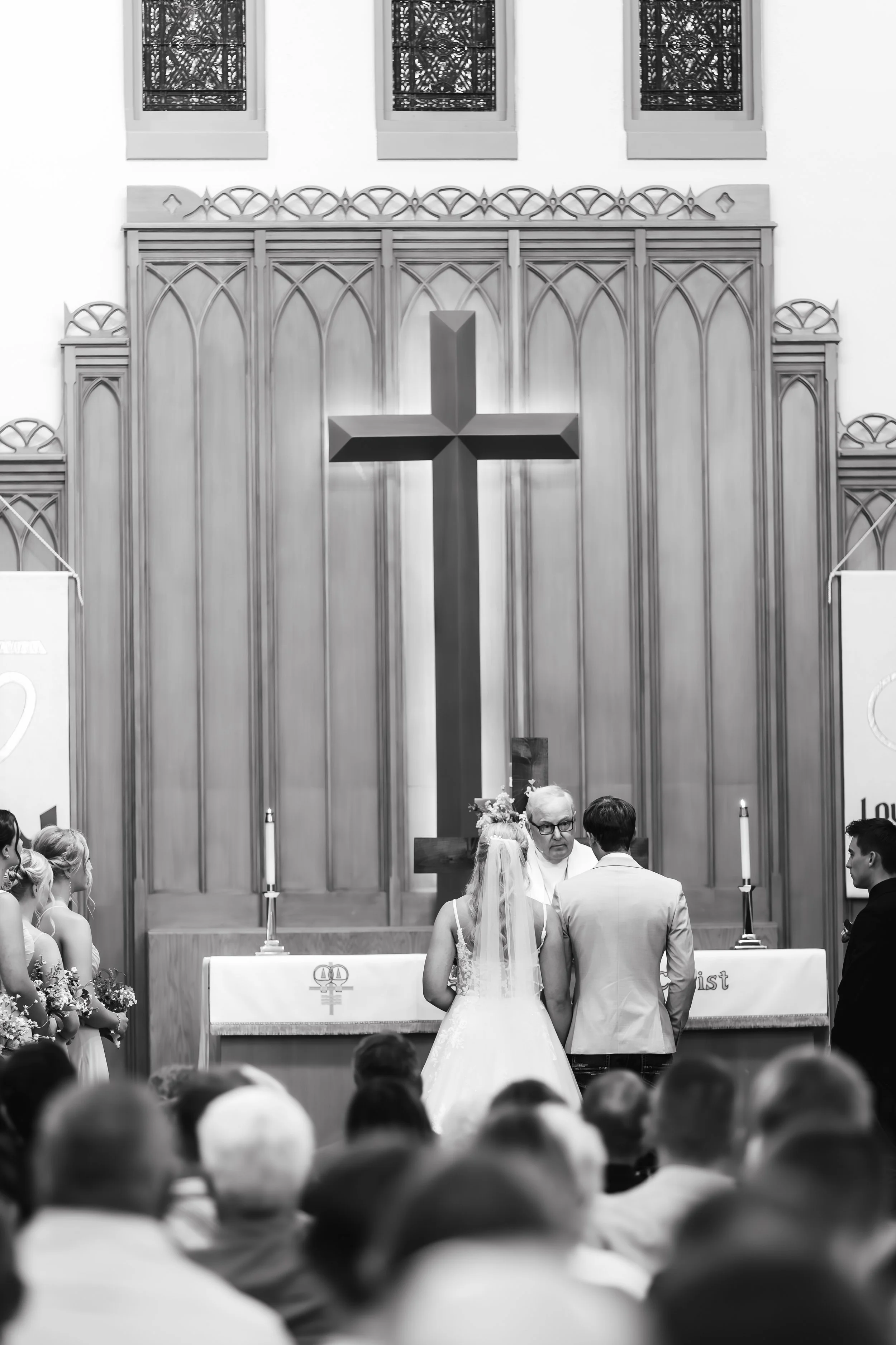 Black and white photo of a wedding ceremony inside a church, with a couple standing before the officiant, facing an altar with a large cross in Luverne, MN.