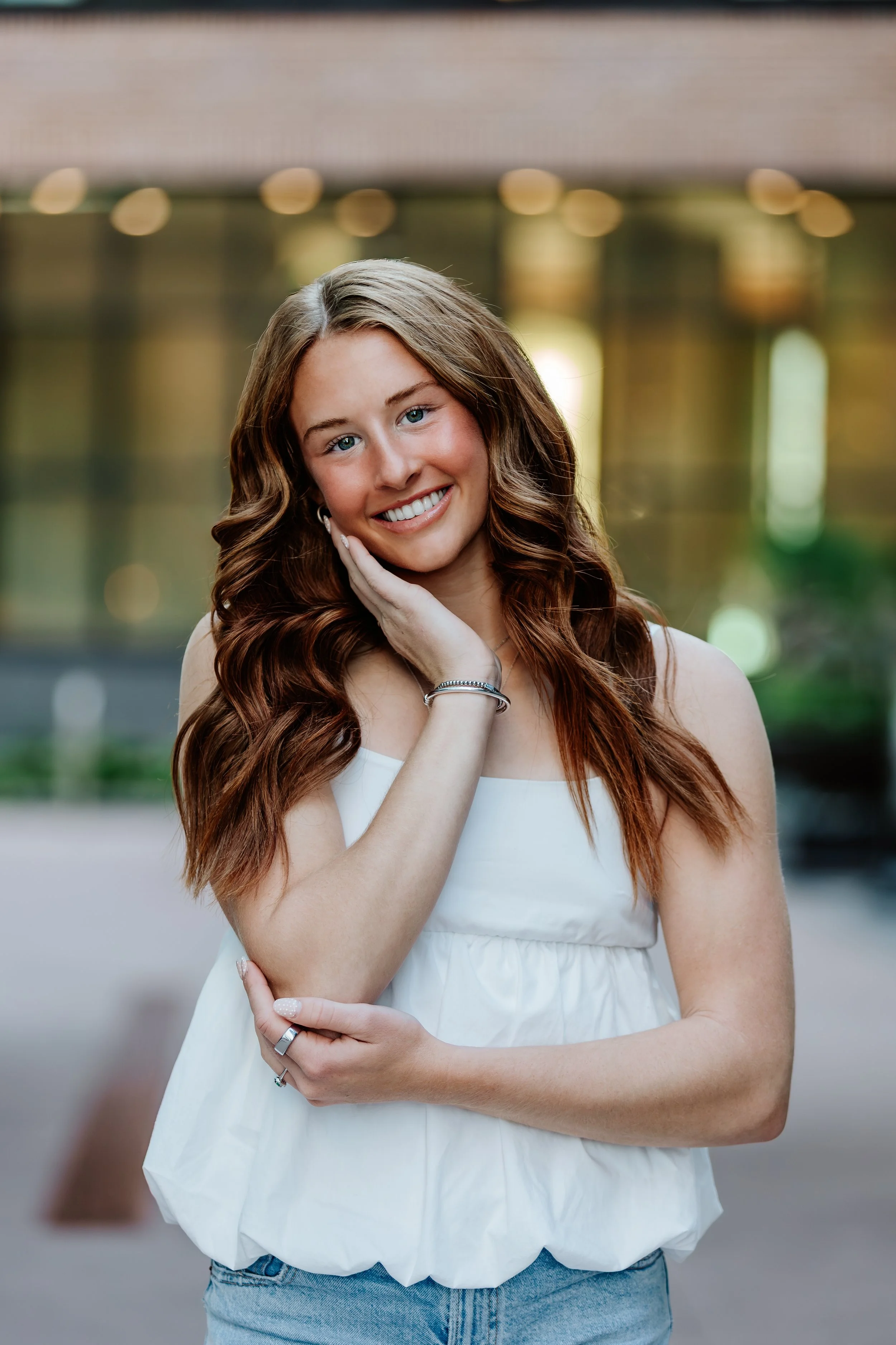 Young woman with long wavy brown hair, smiling, wearing a white sleeveless top and jeans, standing outdoors with a blurred background in Downtown Sioux Falls, South Dakota.