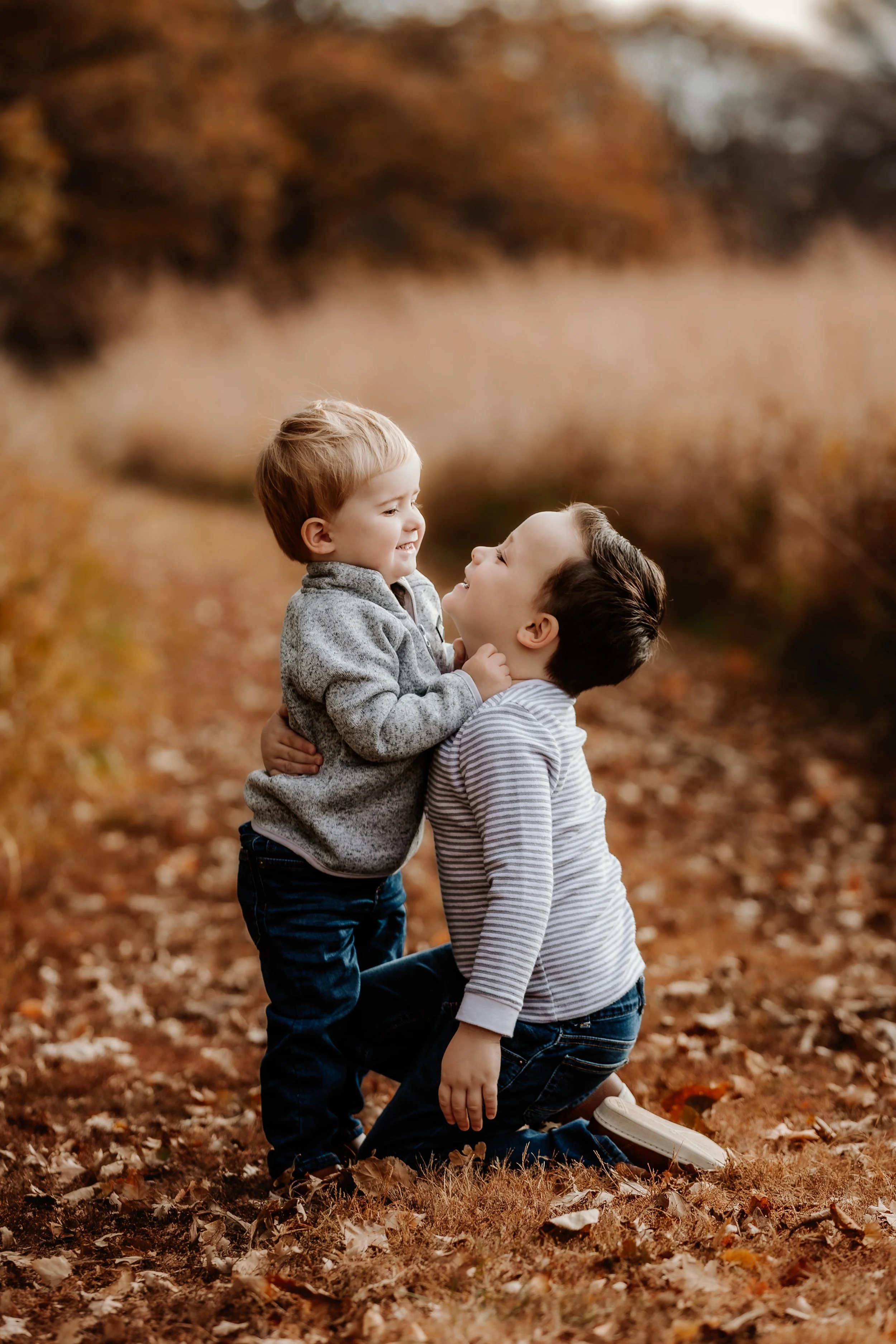 Two young boys playing together outdoors in fall, one kneeling and holding the other at chest level, smiling and looking at each other, with autumn leaves on the ground at Good Earth State Park in Sioux Falls, South Dakota.
