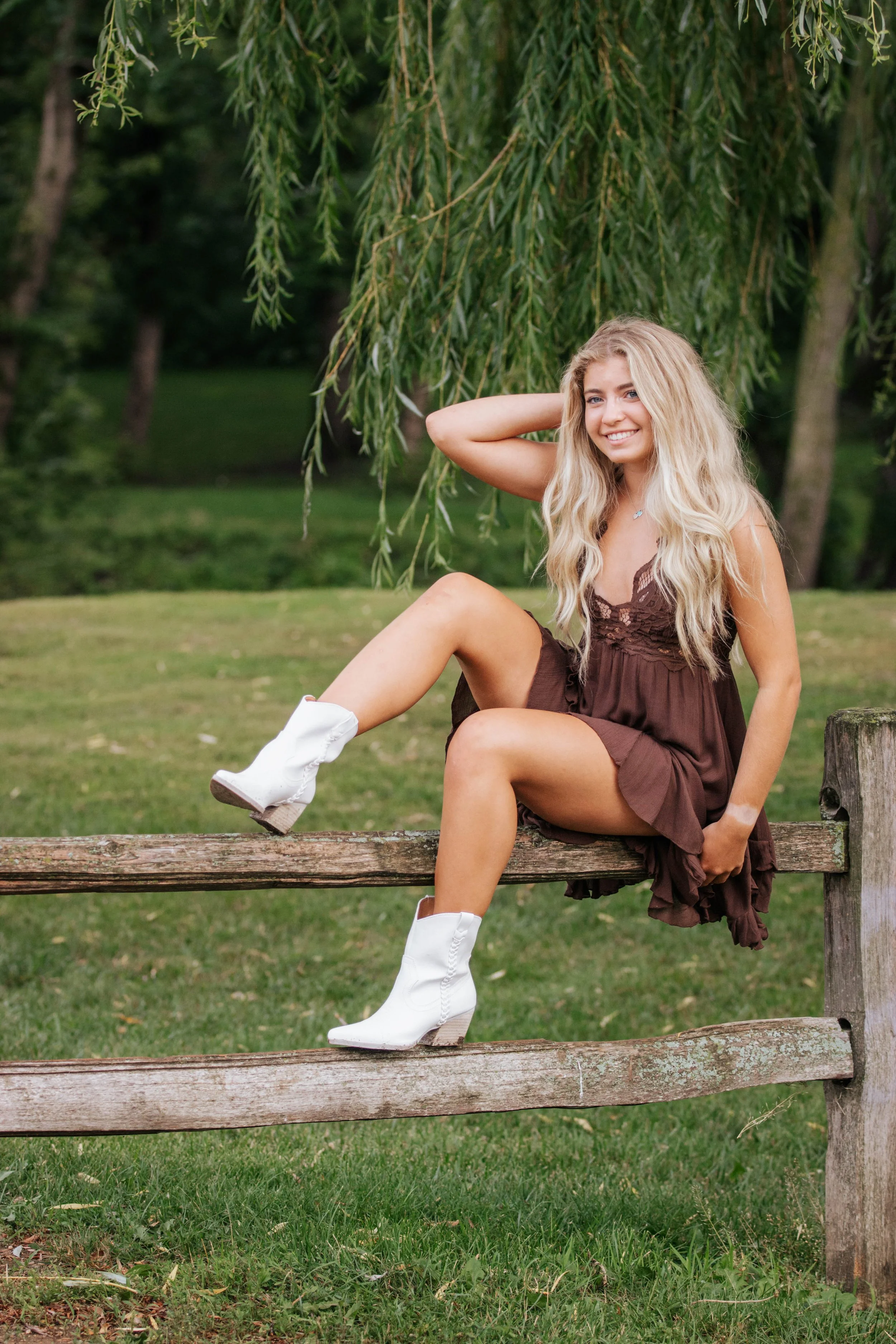 A young woman with long blonde hair, wearing a brown dress and white cowboy boots, sitting on a wooden fence in a park with green grass and trees in Downtown Sioux Falls, South Dakota.