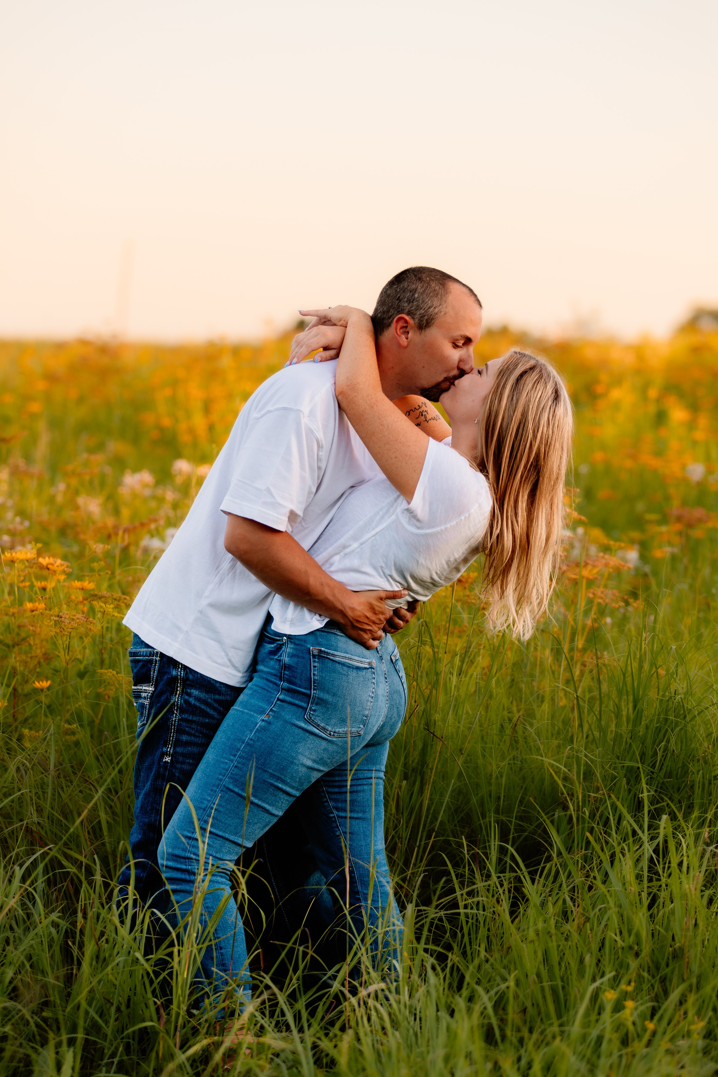 A couple sharing a kiss in a field of yellow flowers during sunset, with the man holding the woman around her waist as they kiss at Good Earth State Park in Sioux Falls, South Dakota.