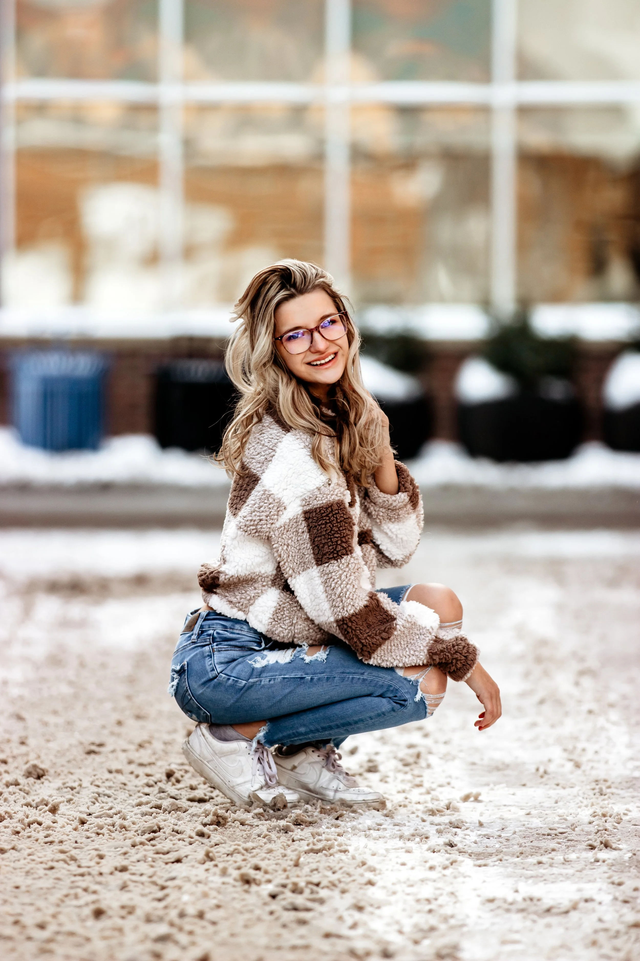 A young woman crouching on a sandy surface outdoors, wearing glasses, a fuzzy checkered jacket, ripped jeans, and sneakers, smiling at the camera against a blurred background in Downtown Sioux Falls, South Dakota.