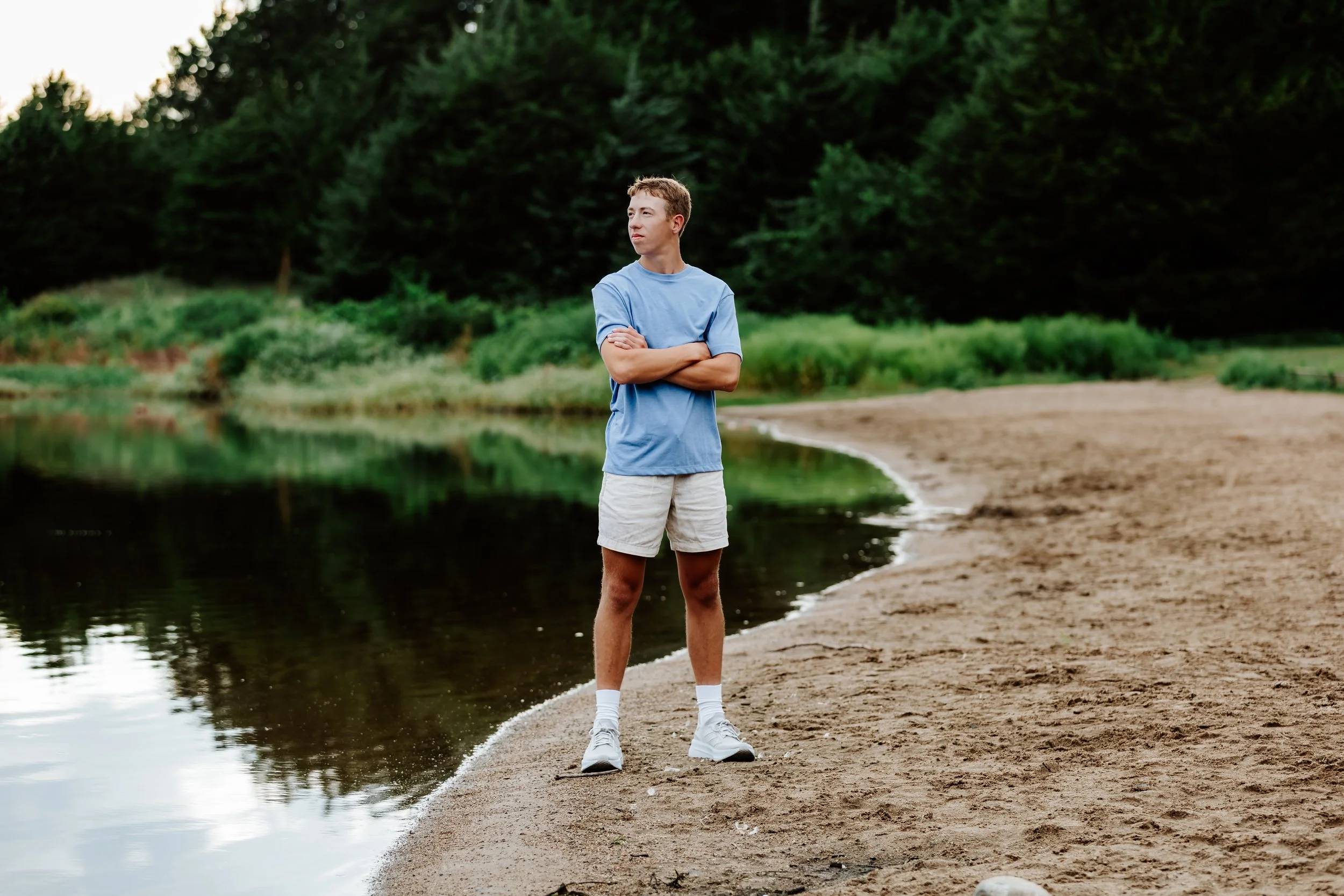 A young man in a blue t-shirt and beige shorts standing with arms crossed on a sandy beach next to a calm body of water, with lush green trees in the background at Lake Alvin in Sioux Falls, South Dakota.