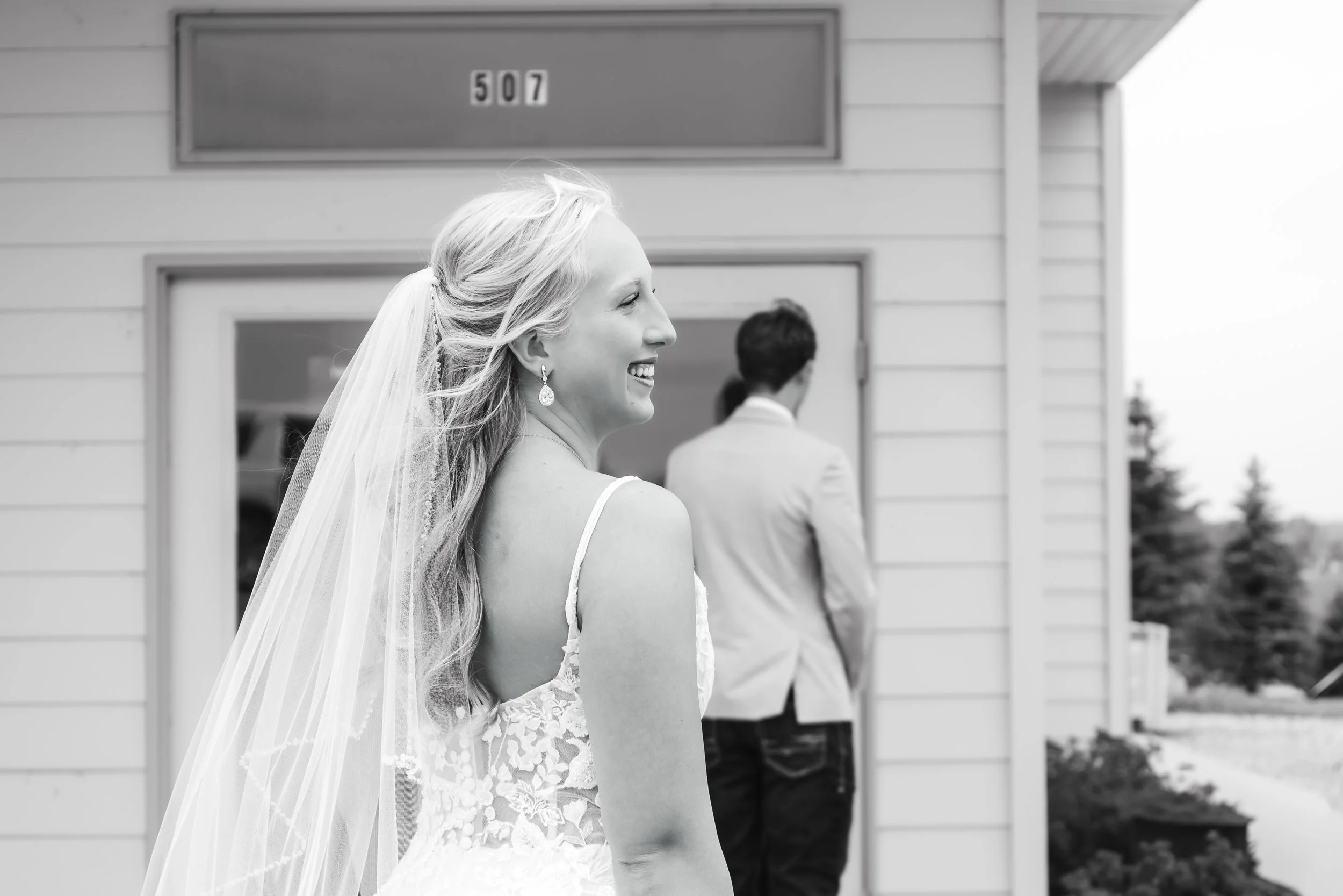 A smiling bride with long, flowing hair and a lace wedding dress stands outside a building, looking to her left, with a man in a suit in the background facing away from the camera in Luverne, MN.