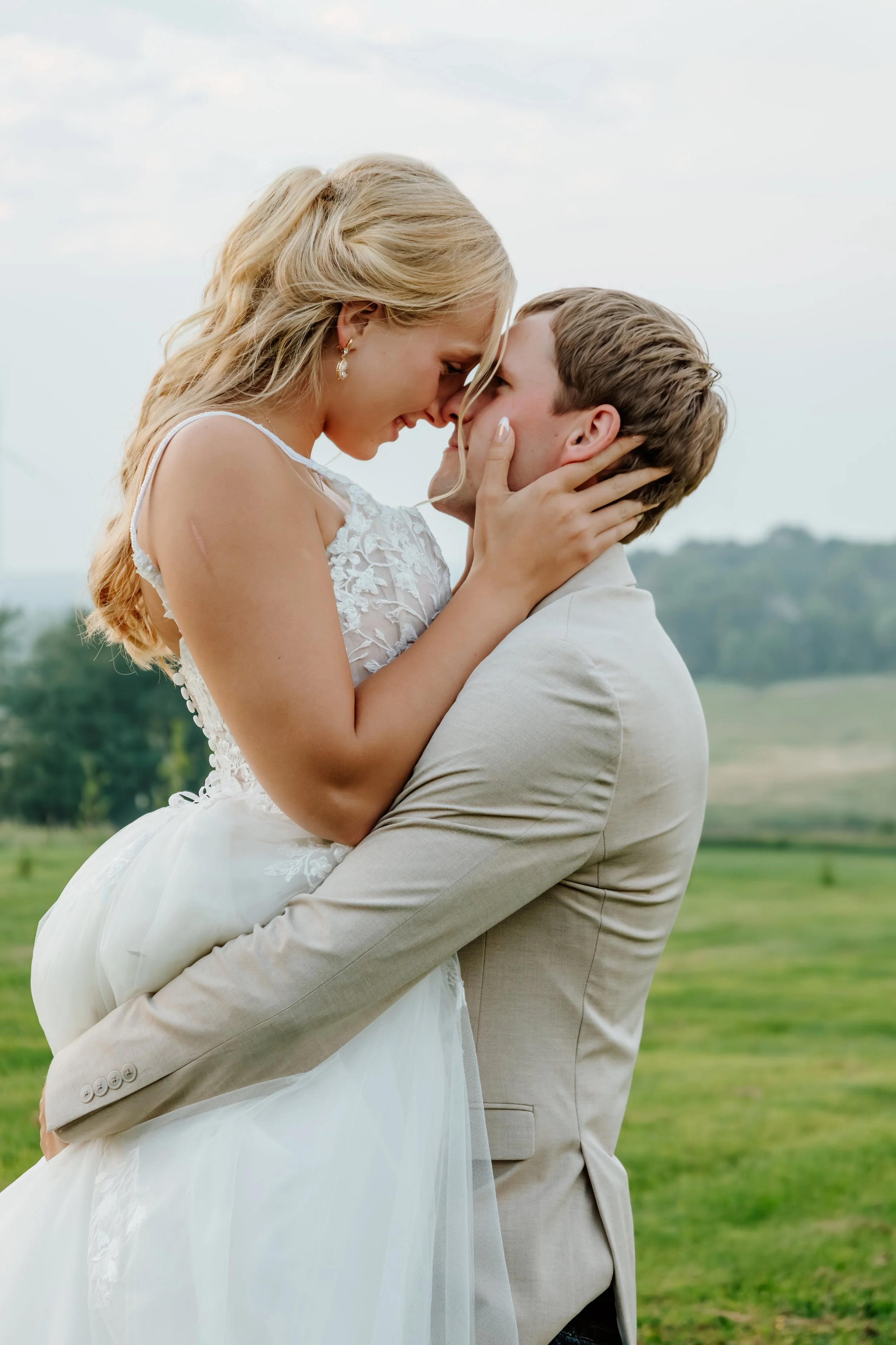 A bride and groom share a romantic moment outdoors during their wedding, with the bride sitting on the groom's lap and both smiling with their foreheads touching in Luverne, MN.