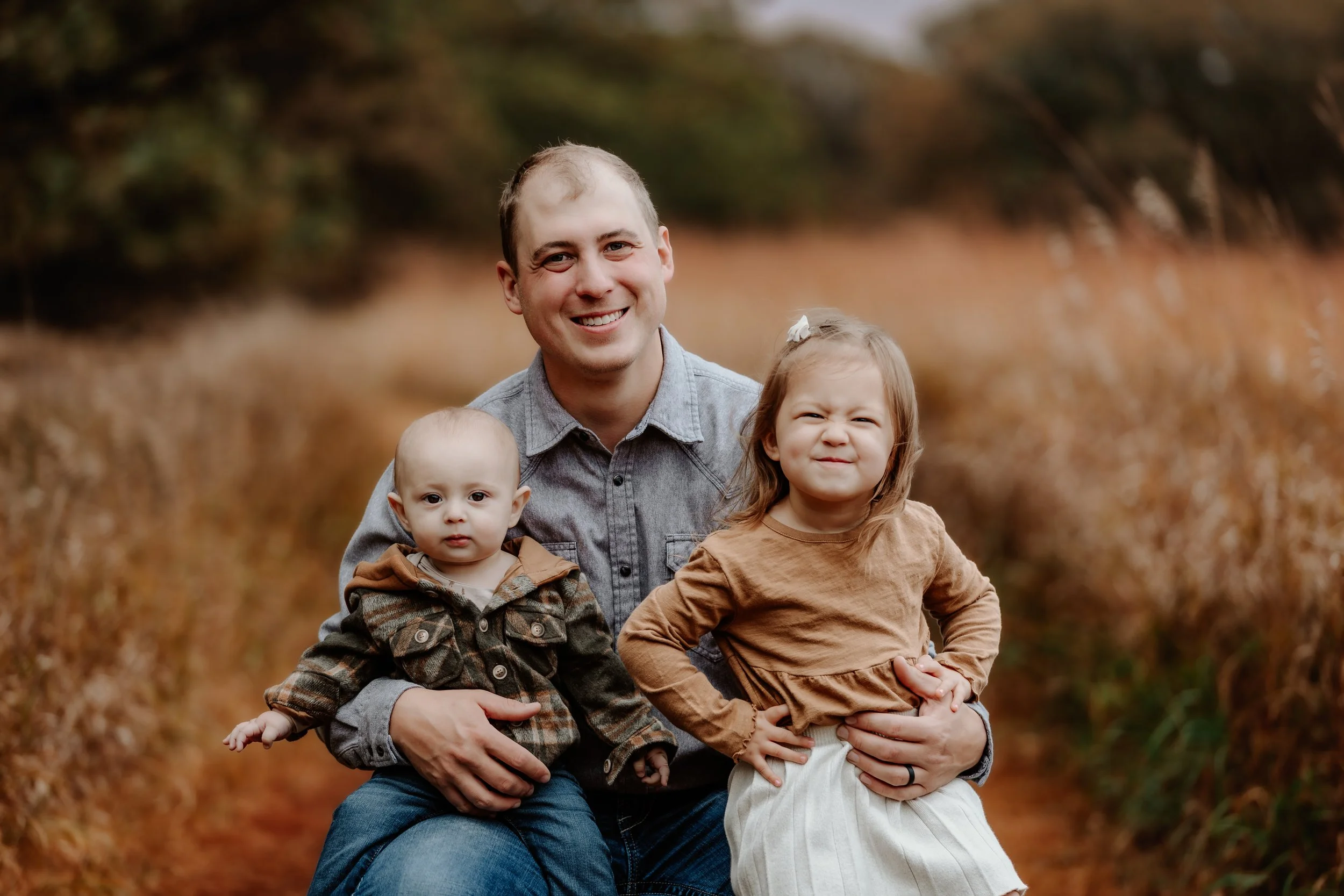 A man smiling outdoors with two children, a baby and a young girl, on an autumn day in a field with brown foliage in Sioux Falls, South Dakota at Good Earth State Park.
