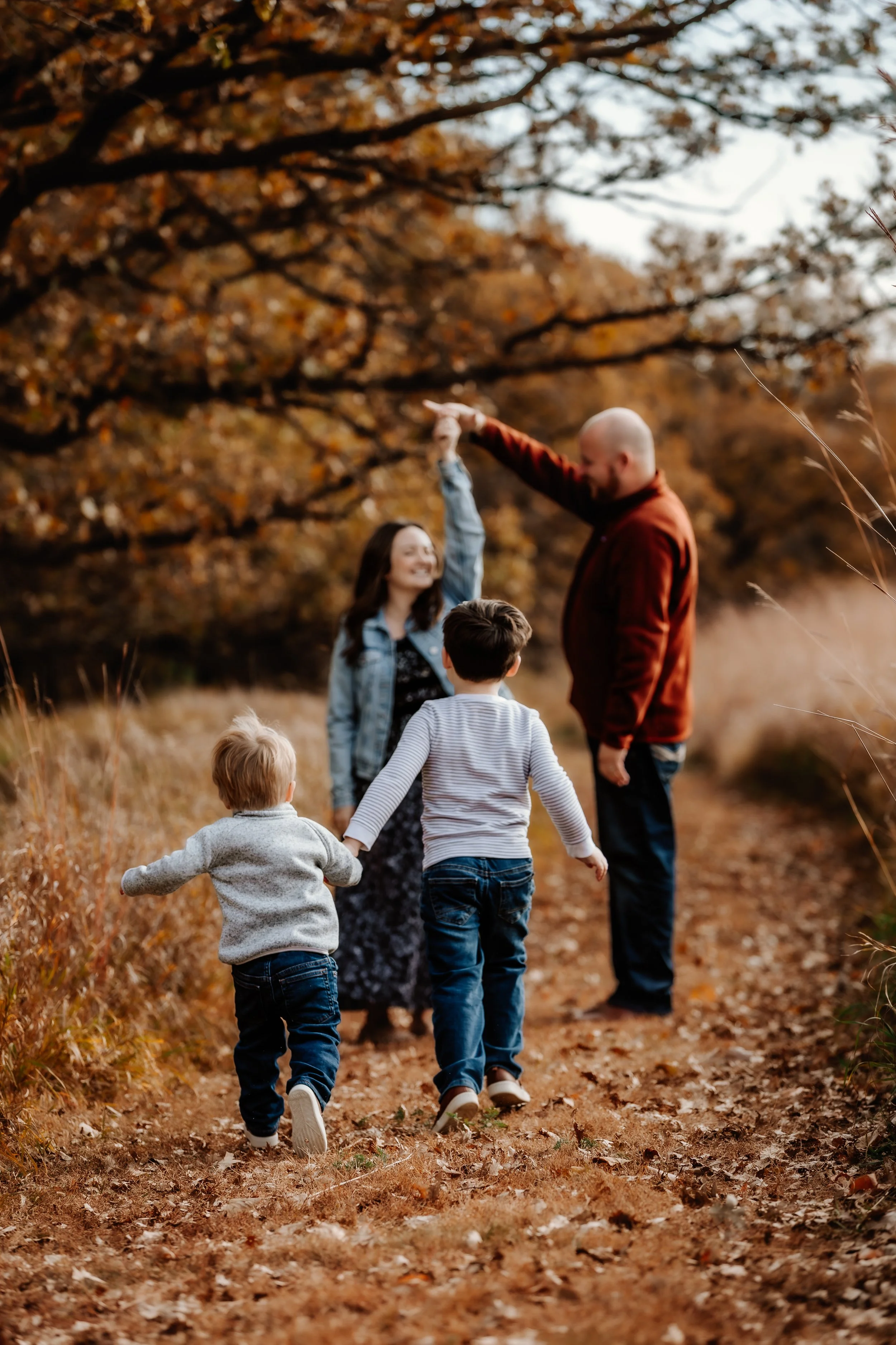 Family of four enjoying a walk on a fall day, holding hands, with autumn trees and fallen leaves in Sioux Falls, South Dakota at Good Earth State Park.