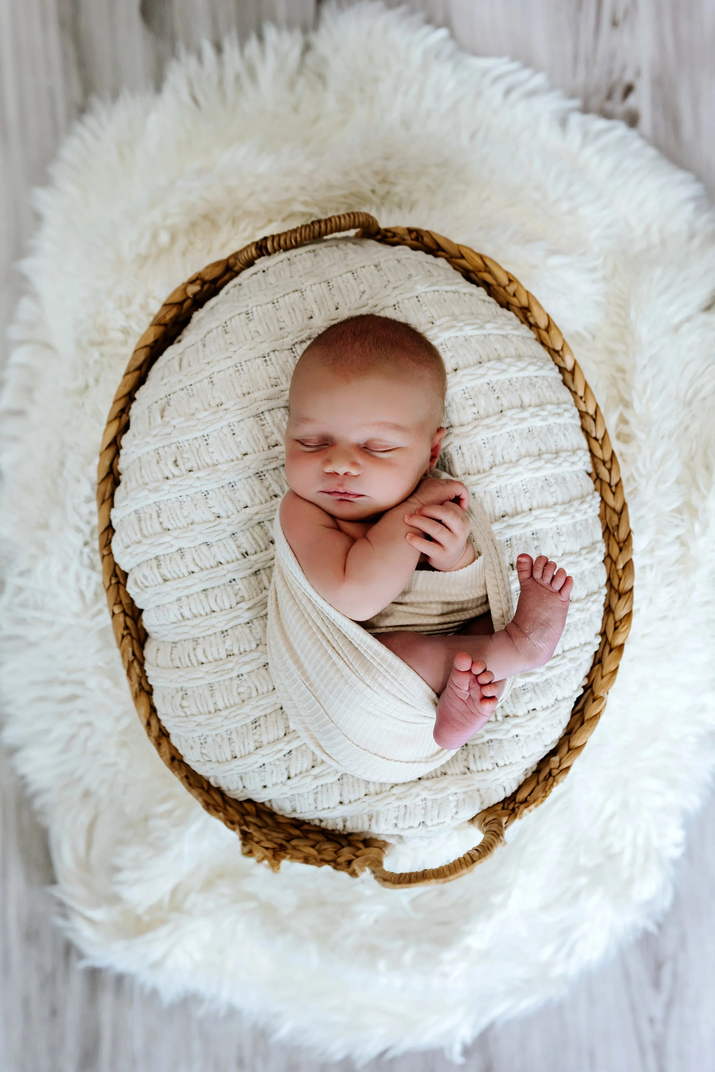 A sleeping baby wrapped in a cream-colored blanket, lying in a woven basket on a fluffy white rug at The White Space Studio in Sioux Falls, South Dakota.