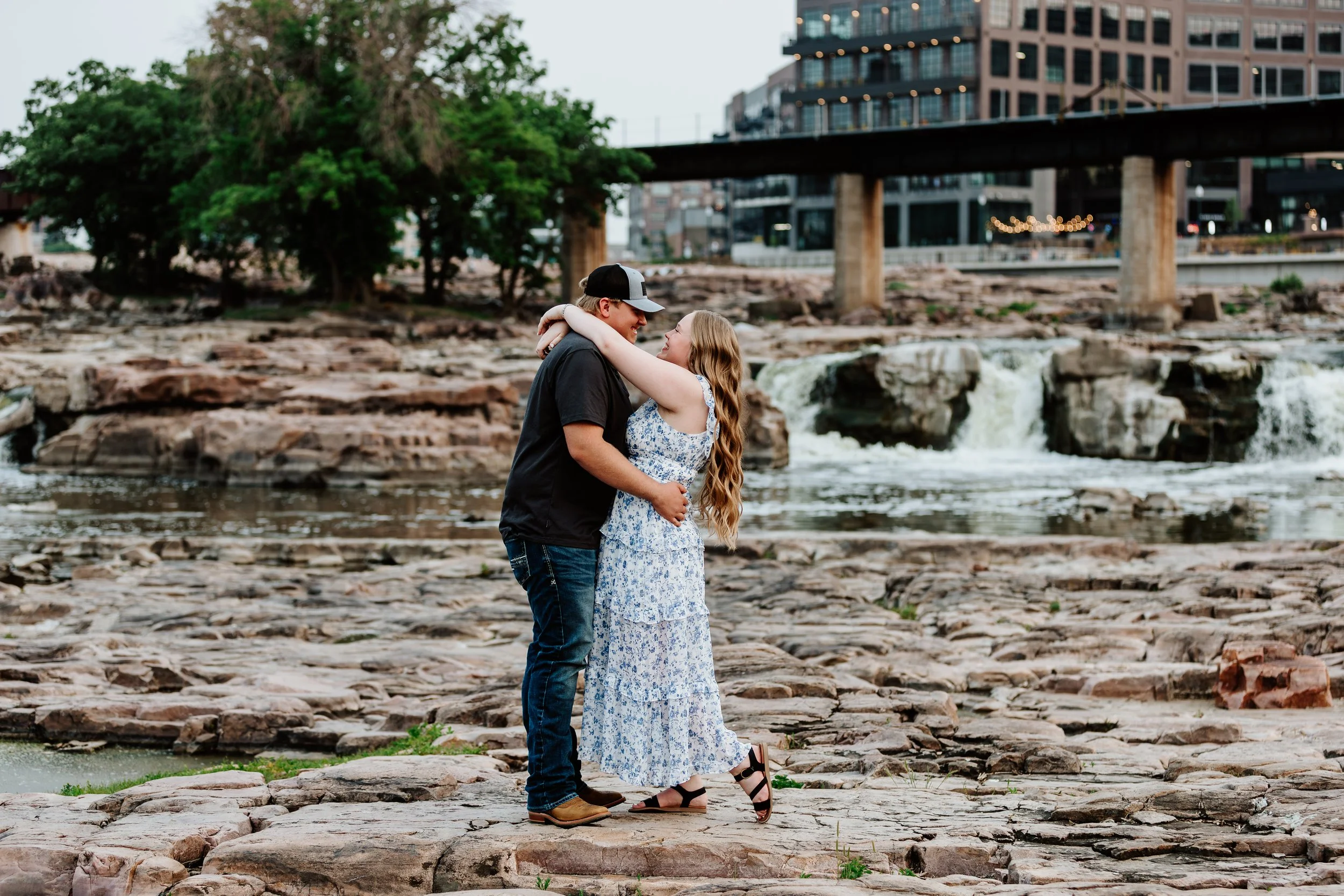 A young couple embraces on rocky riverbank, smiling at each other, with city buildings and a bridge in the background at The Falls Park in Sioux Falls, South Dakota.