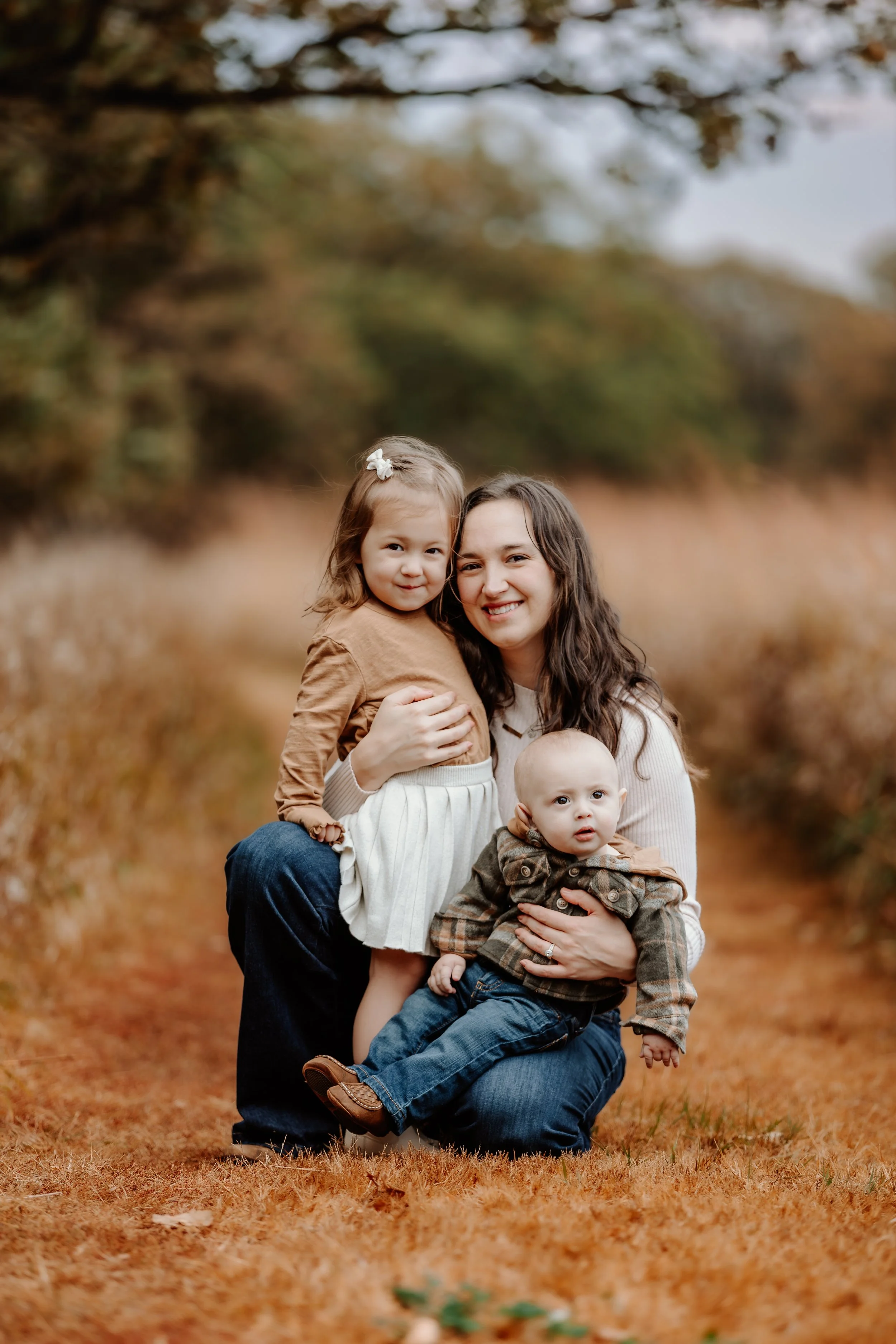 A woman and two young children, a girl and a boy, outdoor in fall attire, smiling and posing for a photo on a leaf-covered path with trees in the background in Sioux Falls, South Dakota at Good Earth State Park.