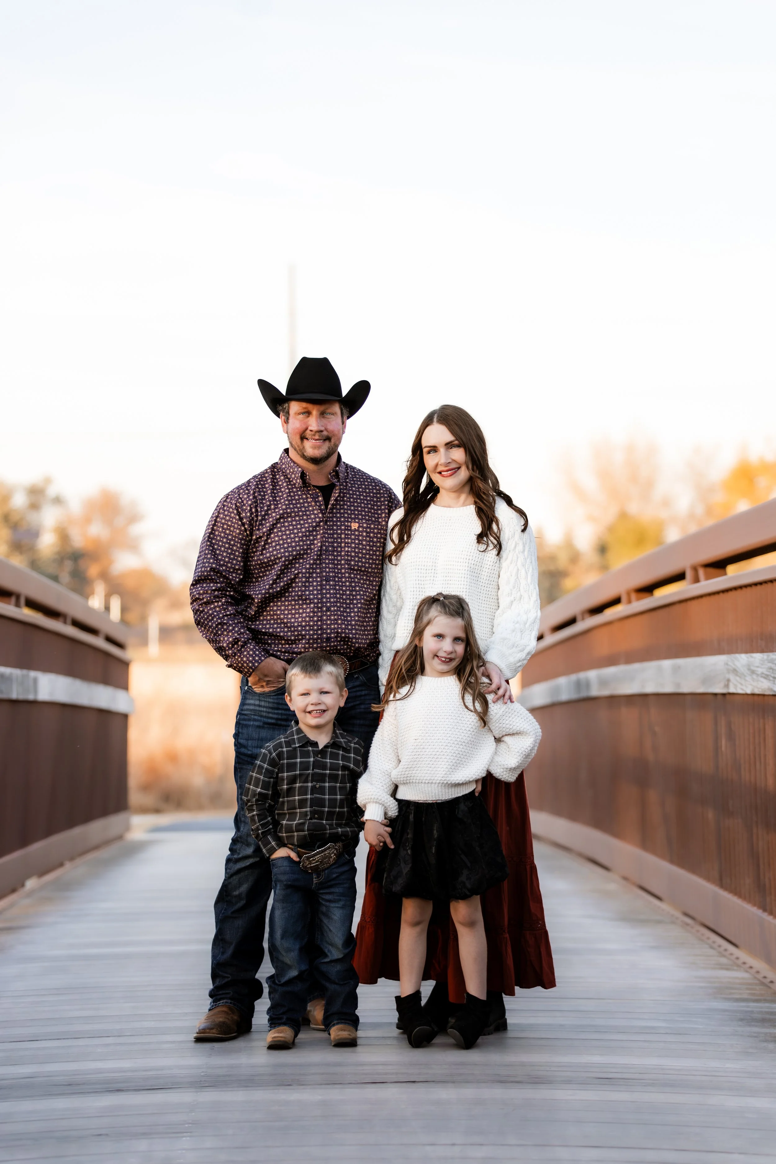 A family of four standing on a wooden bridge outdoors during fall, with trees and a clear sky in the background at Legacy Park in Sioux Falls, South Dakota.