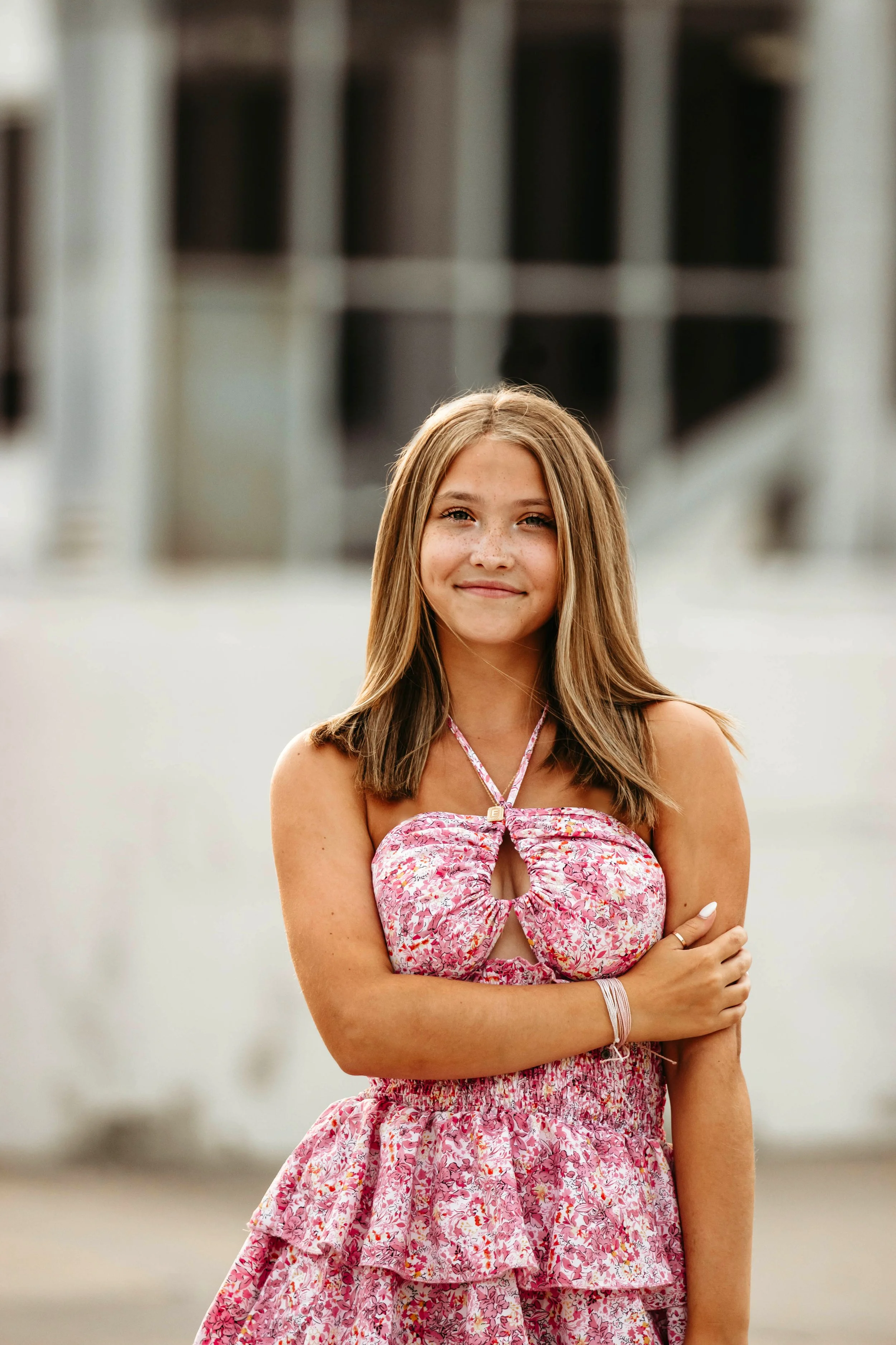 A young girl with light brown hair smiling at the camera, wearing a pink floral dress with a cutout in the front, standing outdoors with a blurred background in Downtown Sioux Falls, South Dakota.