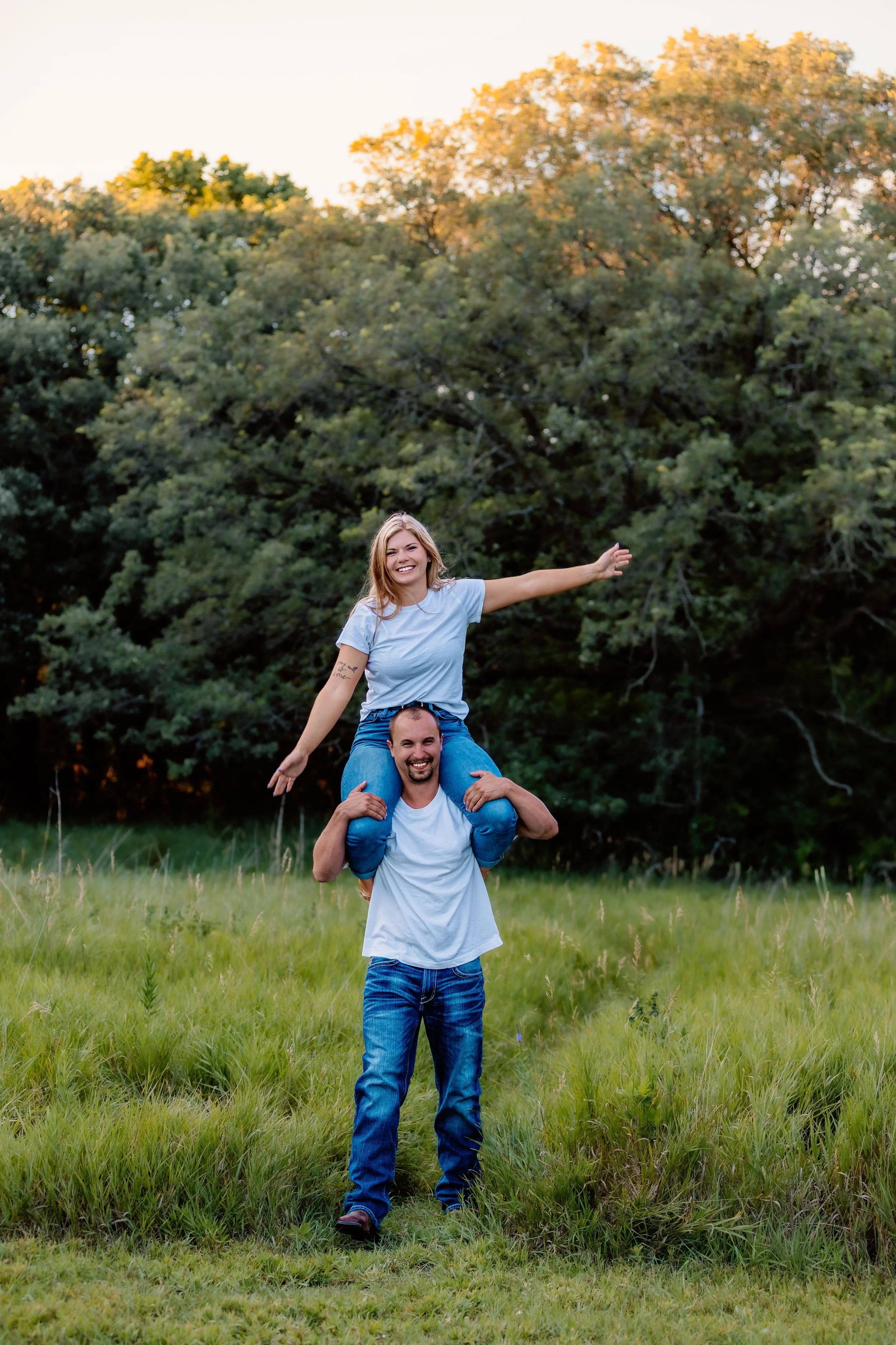A man holding a woman on his shoulders in a grassy field with trees in the background, both smiling at Good Earth State Park in Sioux Falls, South Dakota.