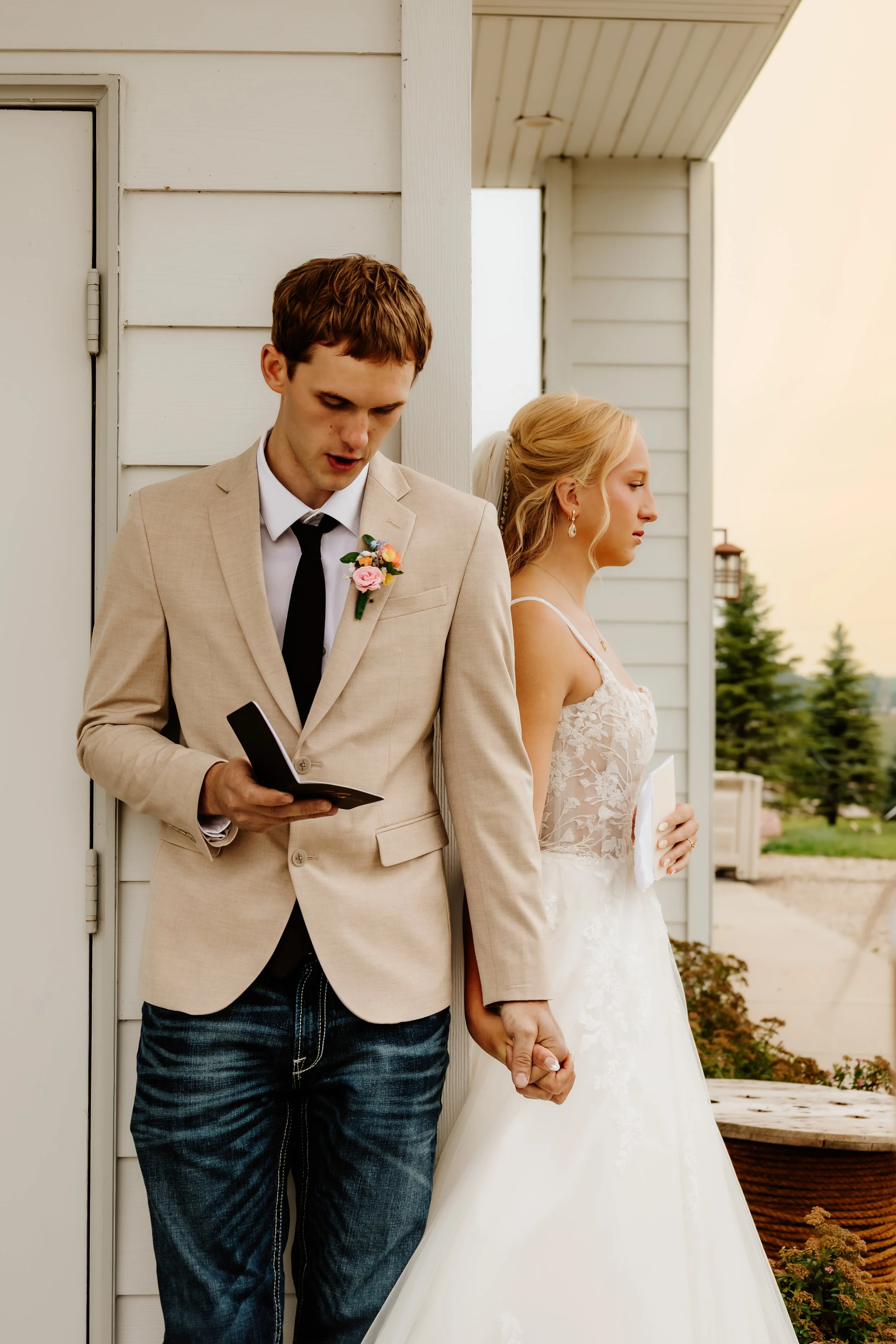 A bride and groom standing back-to-back outside a house, holding hands, with the groom reading from a booklet or paper in Luverne, MN.