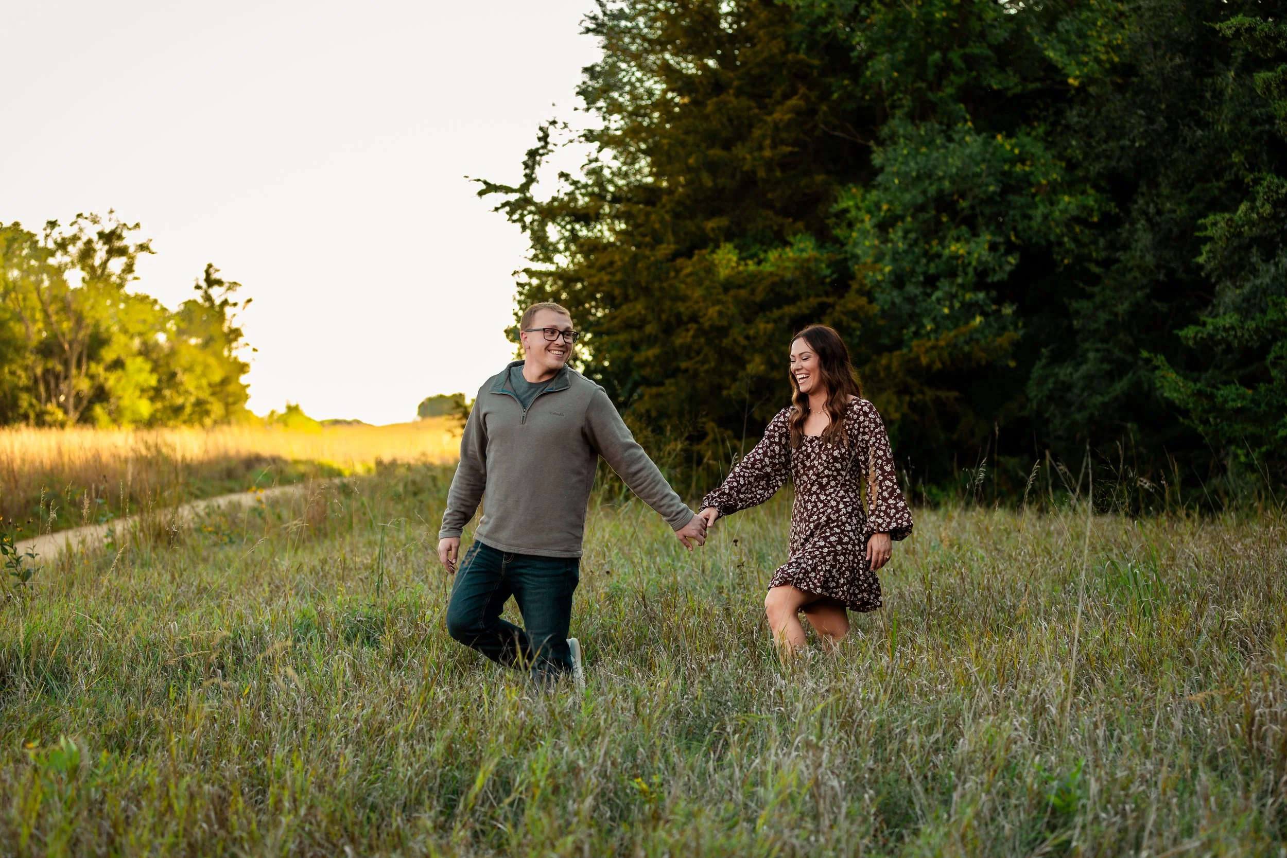 A happy couple holding hands and walking through a grassy field with trees in the background during sunset at Good Earth State Park in Sioux Falls, South Dakota.