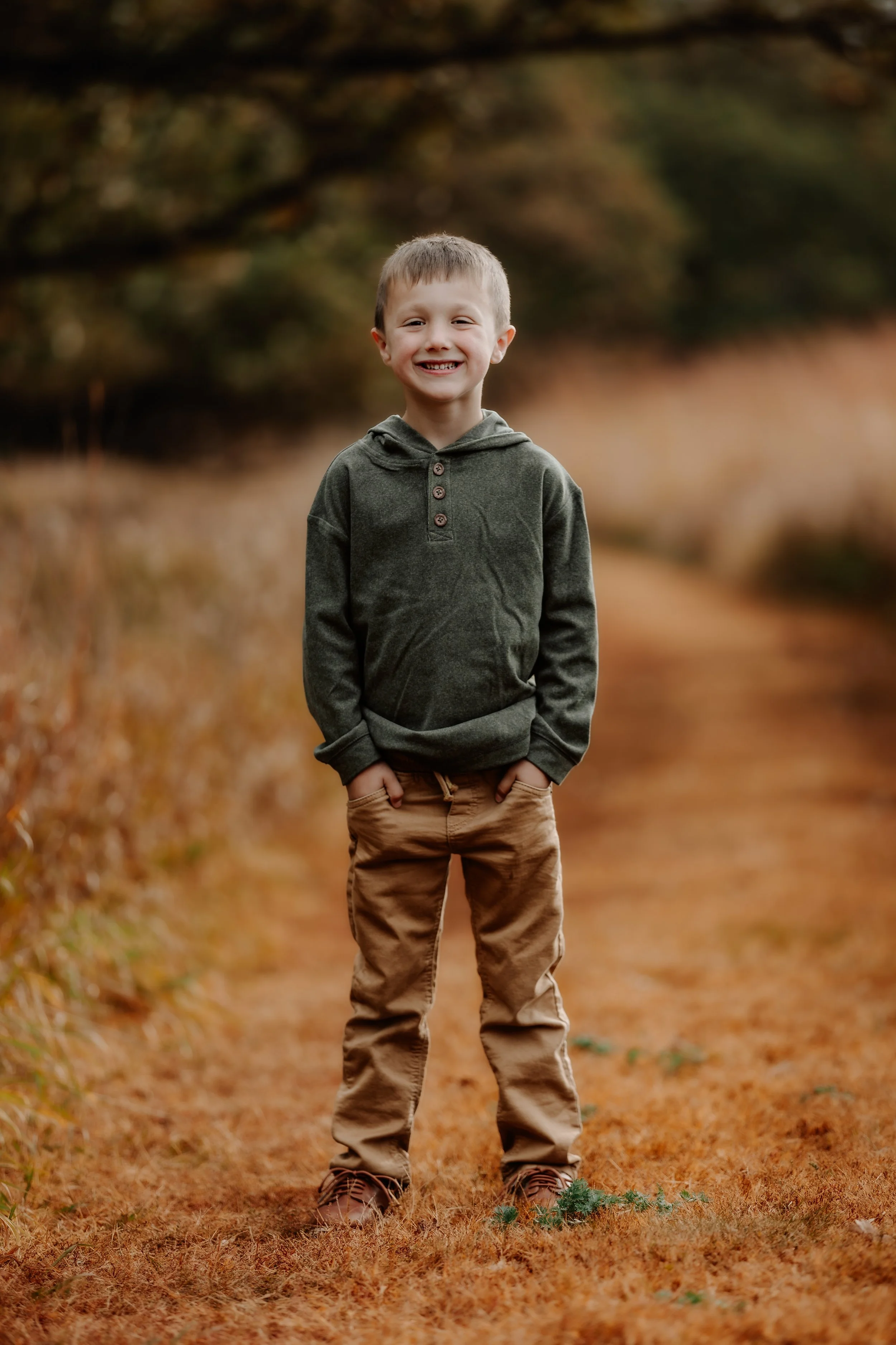 A young boy smiling and standing with hands in pockets on a fall trail with trees and autumn leaves in the background in Sioux Falls, South Dakota at Good Earth State Park.