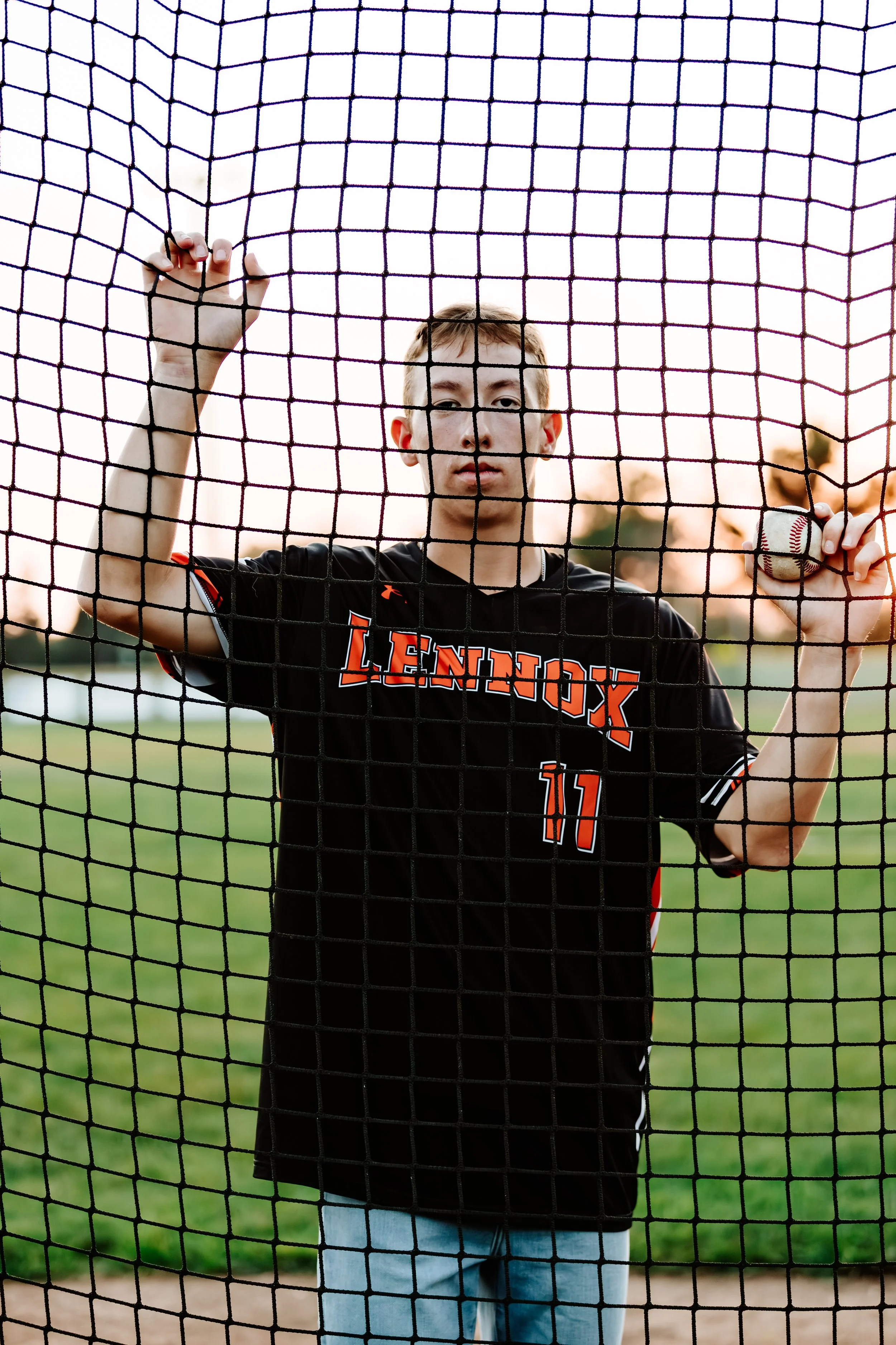 A young male baseball player is standing behind a protective net, holding a baseball and a bat, wearing a black jersey in Lennox, South Dakota.