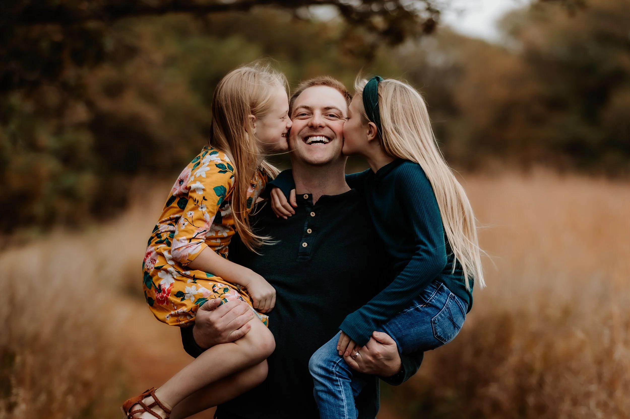 A man holding two young girls, one on each arm, outdoors in a field with autumn trees, both girls kissing the man's cheeks at Good Earth State Park in Sioux Falls, South Dakota.
