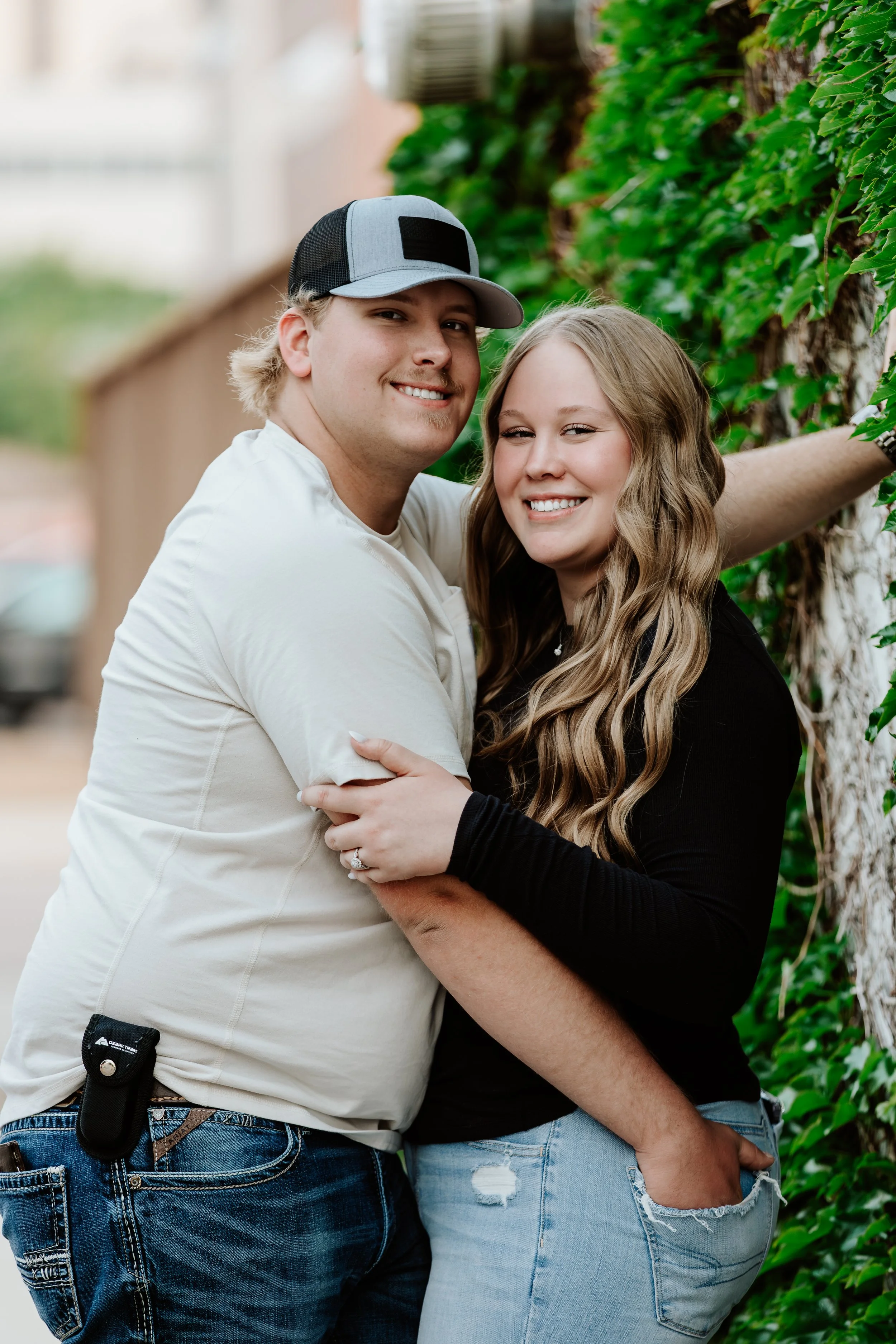 A smiling couple embracing outdoors near a green ivy-covered wall.