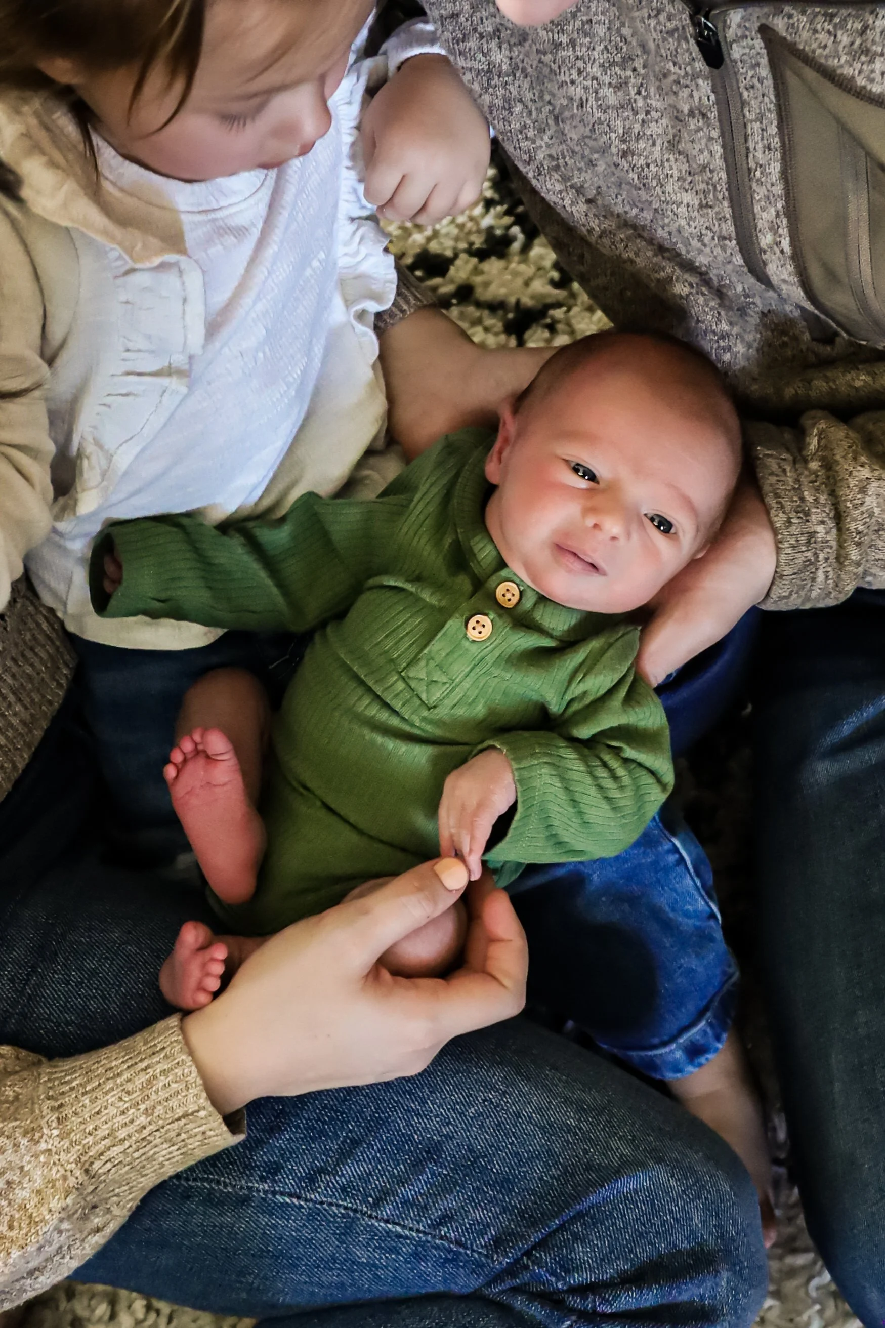 A baby with a green sweater lying on someone's lap, being gently touched on the foot by an adult's hand, surrounded by two children and adult arms in Sioux Falls, South Dakota.