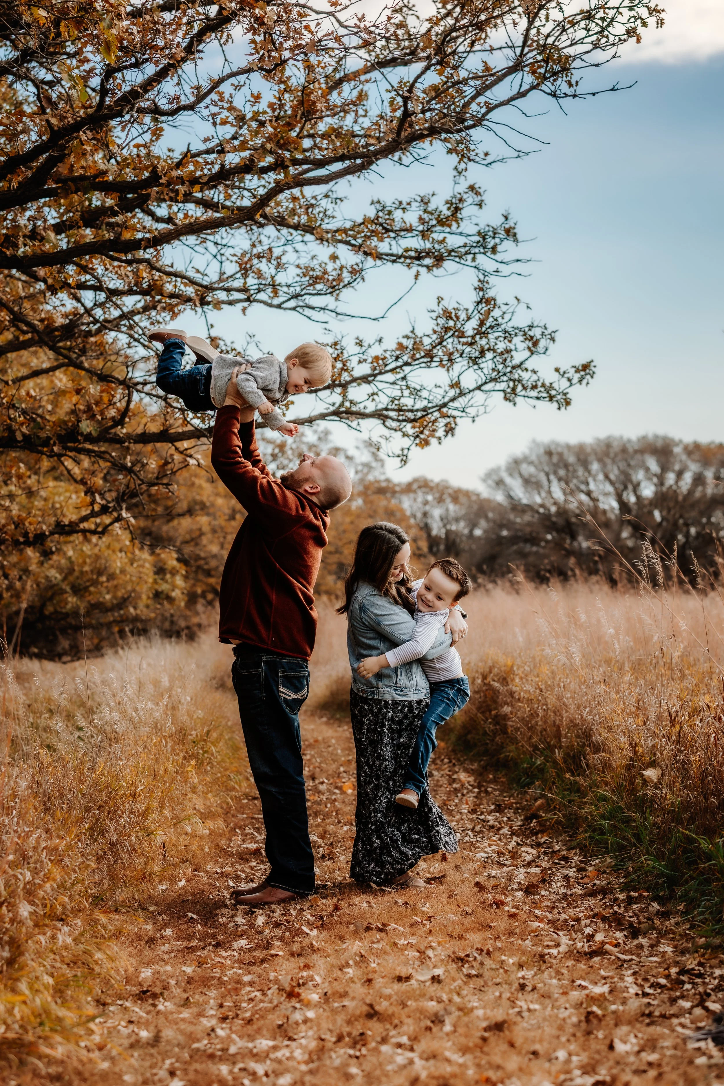 A family of four enjoying fall outdoors, with parents lifting their children up on a trail with autumn foliage in the background in Sioux Falls, South Dakota at Good Earth State Park.