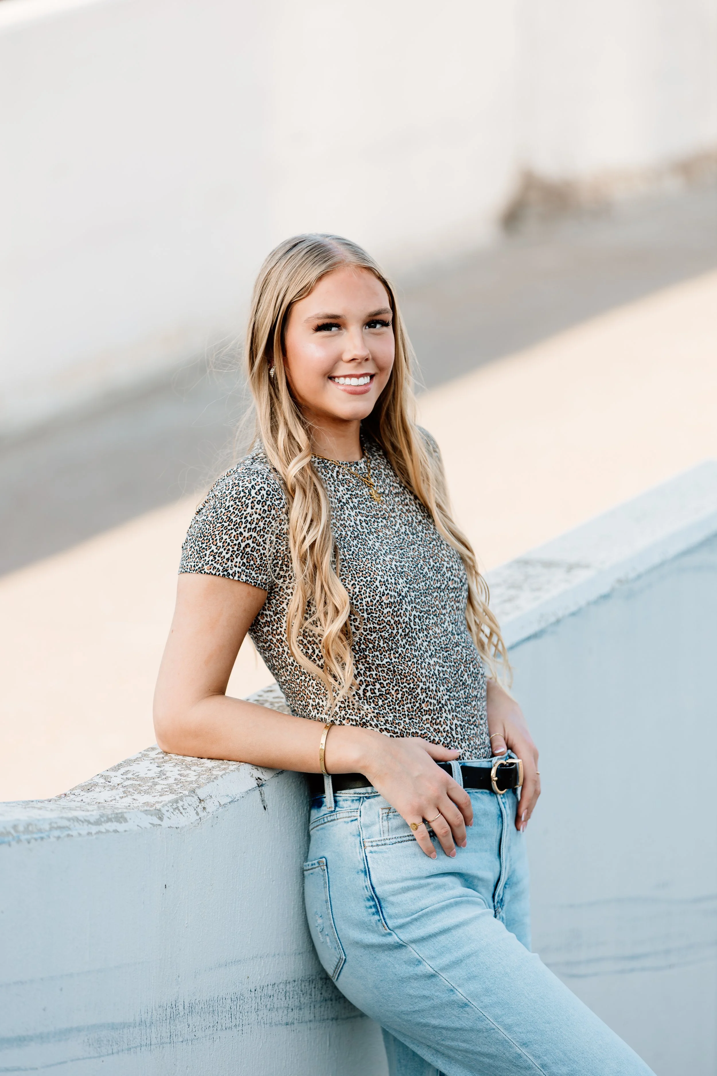 Young woman with long blonde hair, smiling, leaning against a white wall, wearing a leopard print top and light blue jeans in Downtown Sioux Falls, South Dakota.