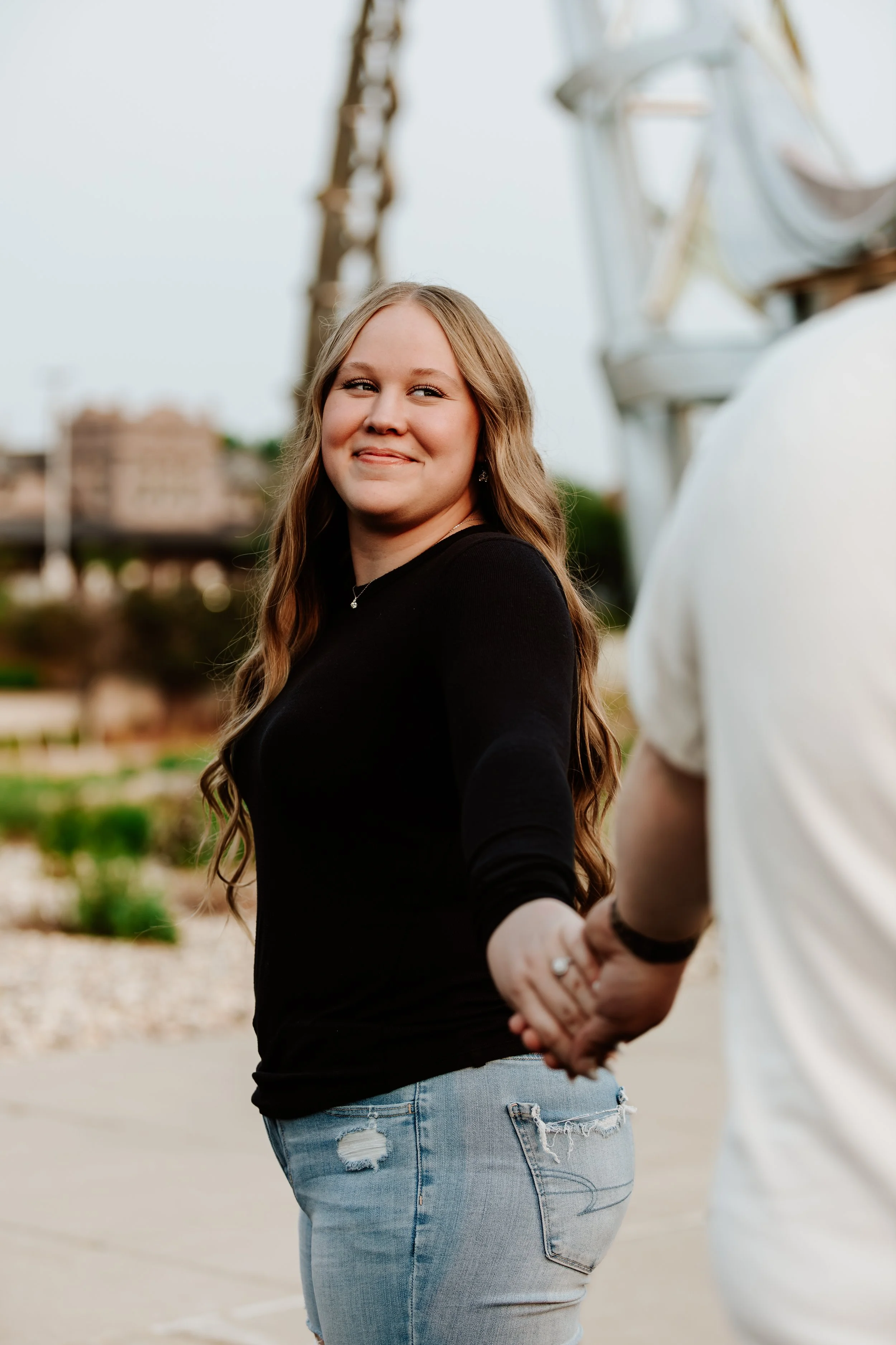 A smiling young woman with long wavy hair holding hands with someone outside, with a Ferris wheel and buildings in the background in Downtown Sioux Falls, South Dakota.