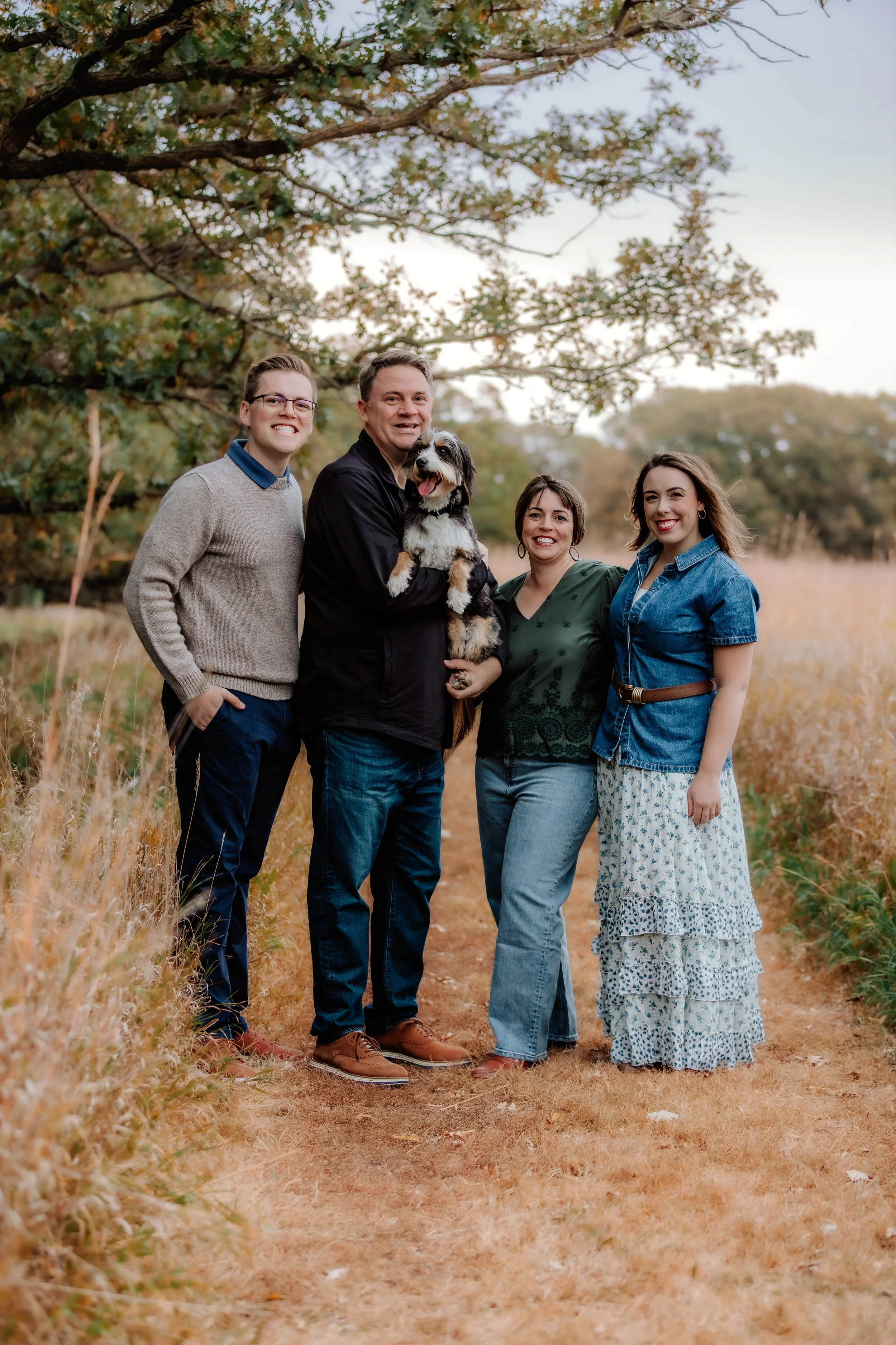 A family of four with a dog standing outdoors on a dirt path surrounded by grass and trees, smiling at the camera in Sioux Falls, South Dakota at Good Earth State Park.