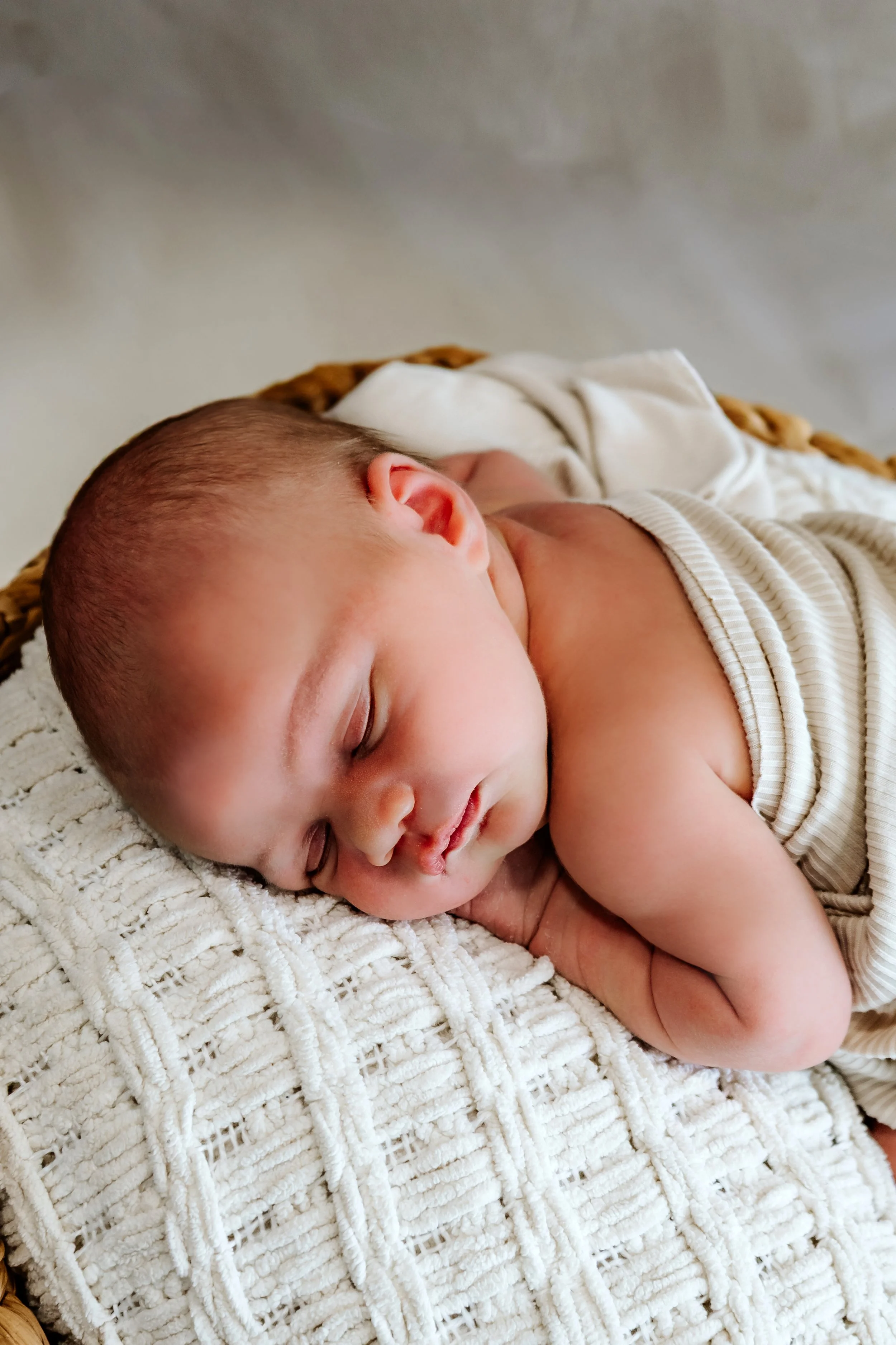 A sleeping baby with a round face and soft skin, resting on a textured white blanket with a wicker basket underneath at The White Space Studio in Sioux Falls, South Dakota.