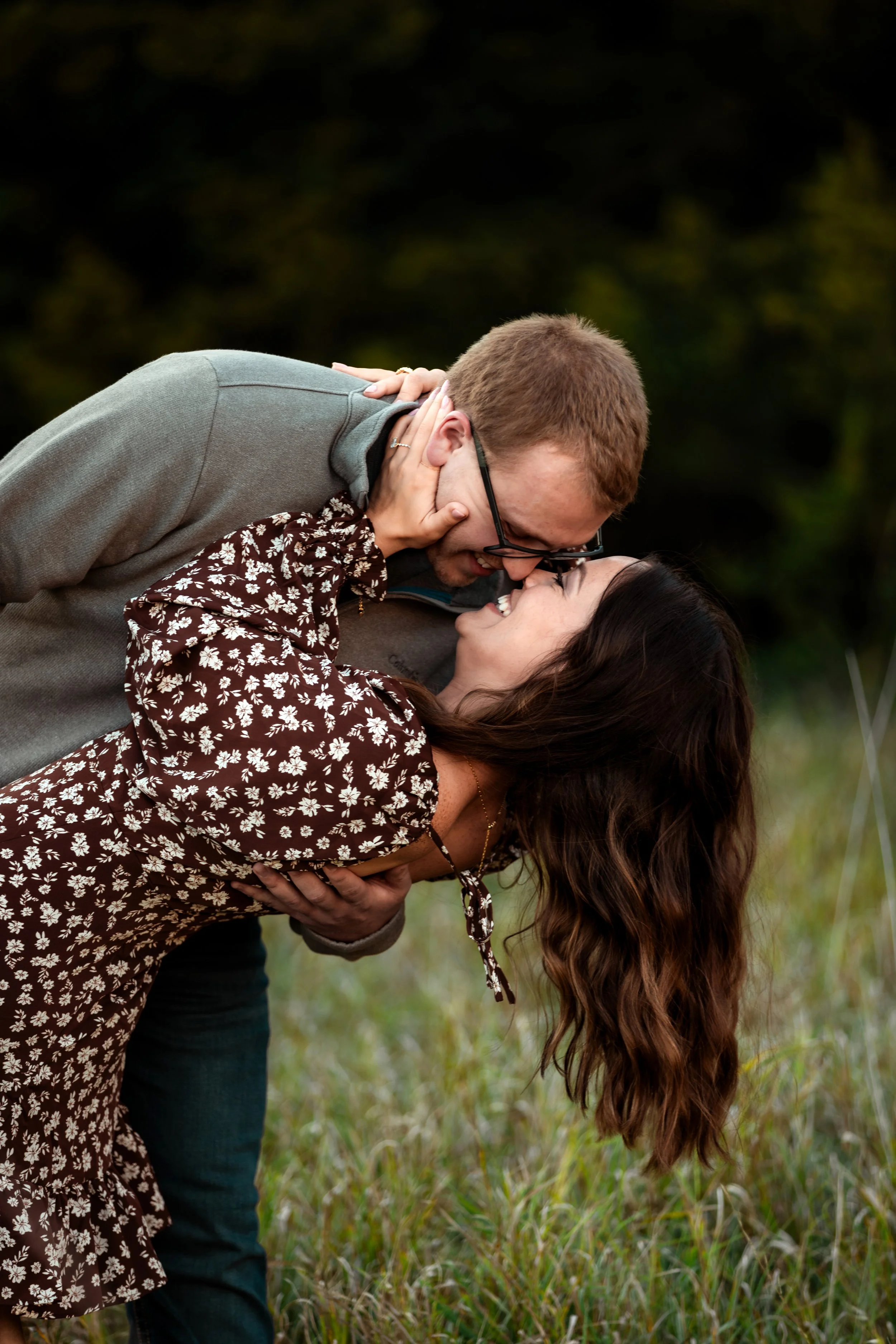 A couple sharing a kiss outdoors in a grassy area with a blurred background of trees at Good Earth State Park in Sioux Falls, South Dakota.