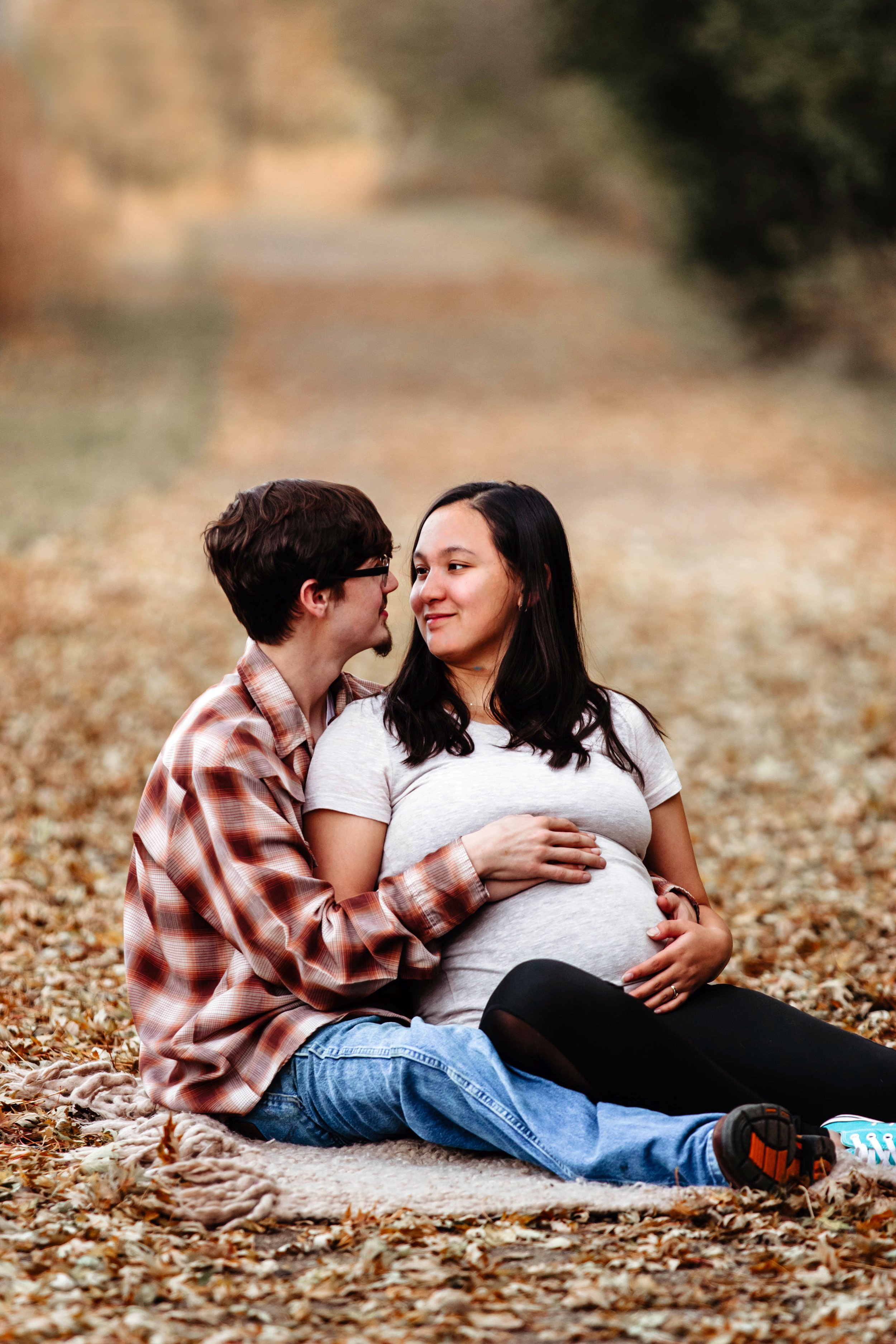 A pregnant woman and a man sitting together on a blanket in a park with fallen leaves, facing each other happily in Sioux Falls, South Dakota at The outdoor Campus.