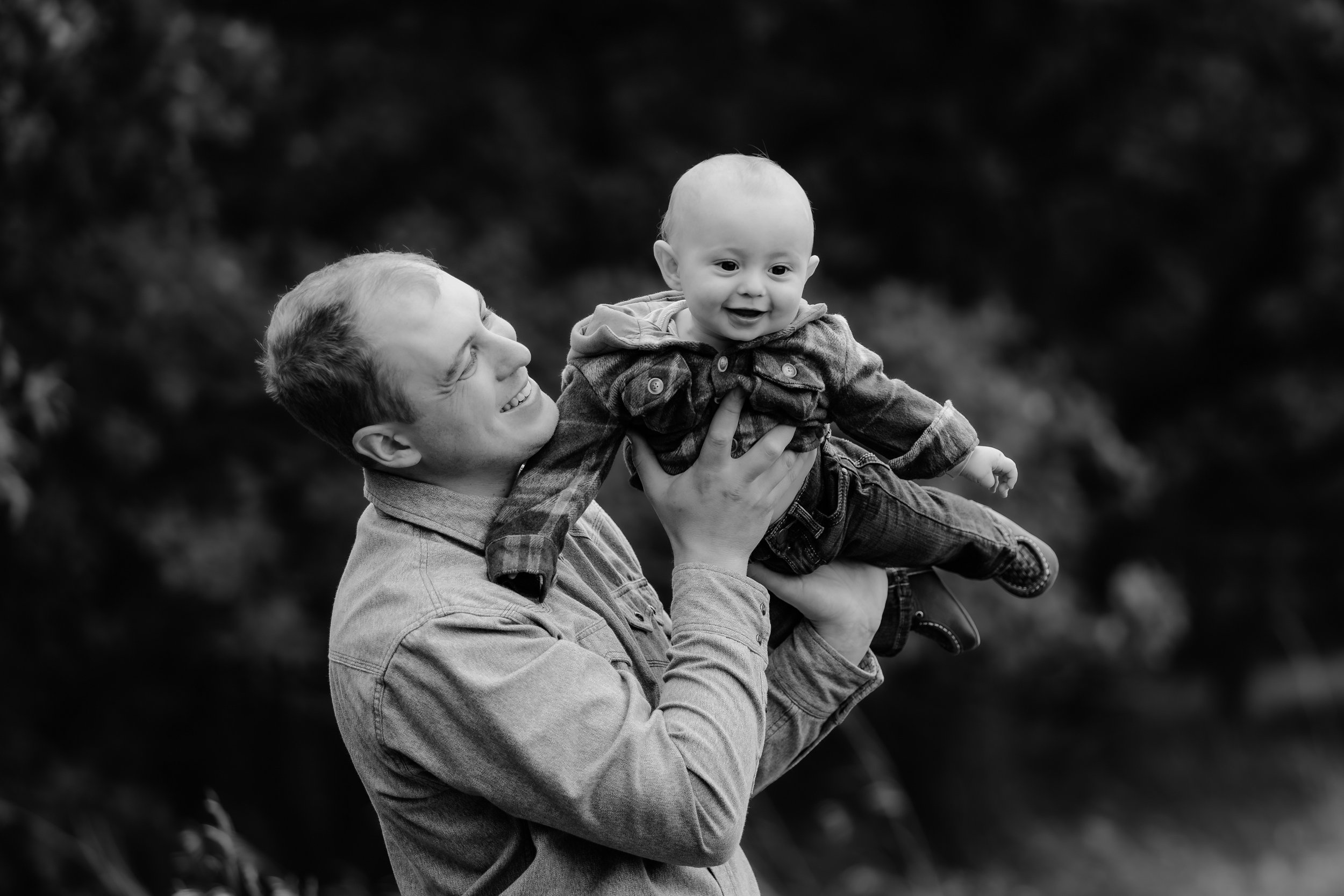 A man is lifting a young child in the air outside. The man is smiling at the child, who is also smiling. The background is blurred with trees in Sioux Falls, South Dakota at Good Earth State Park.