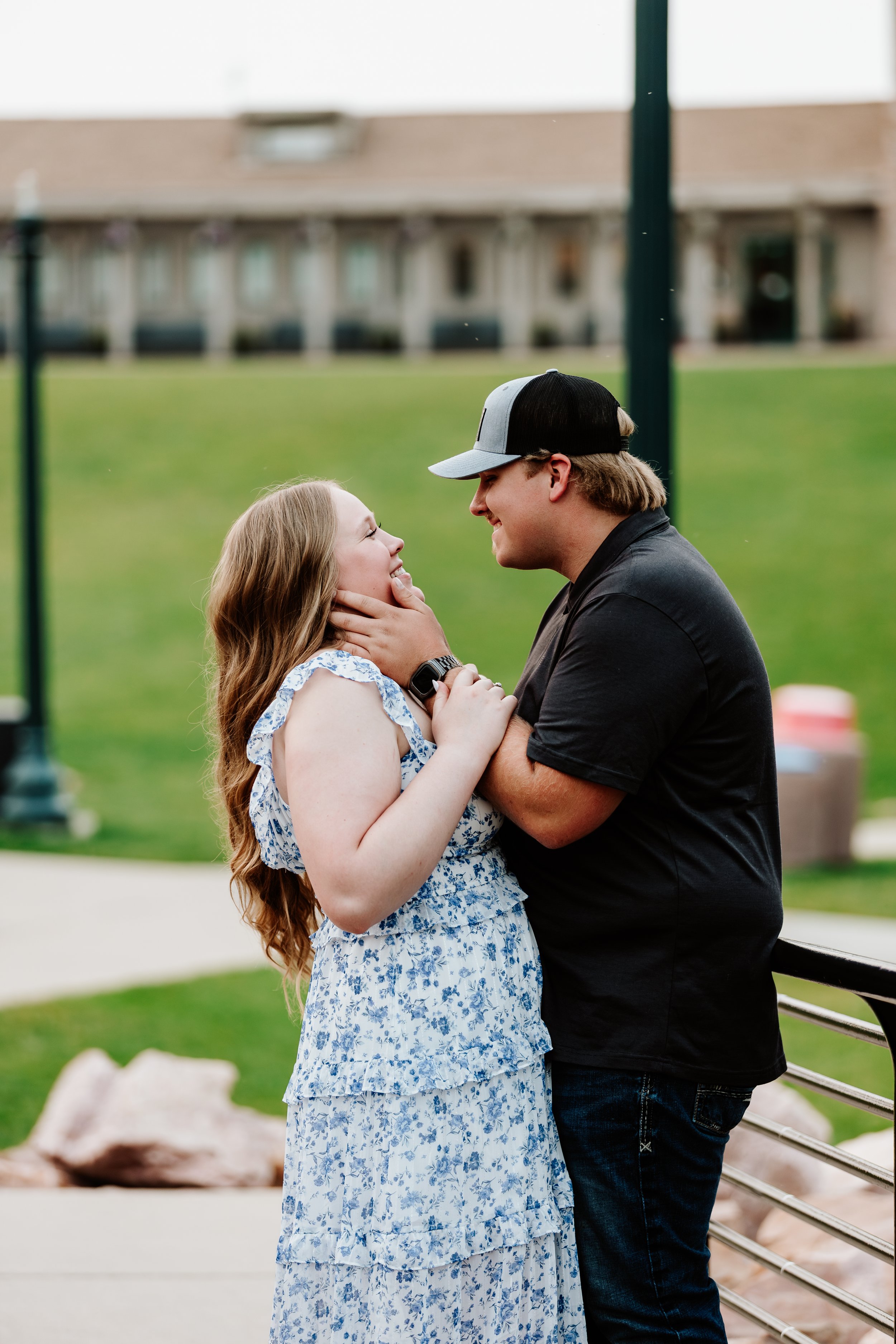A couple smiling at each other outdoors near a bench, with a grassy background and a building in the distance.