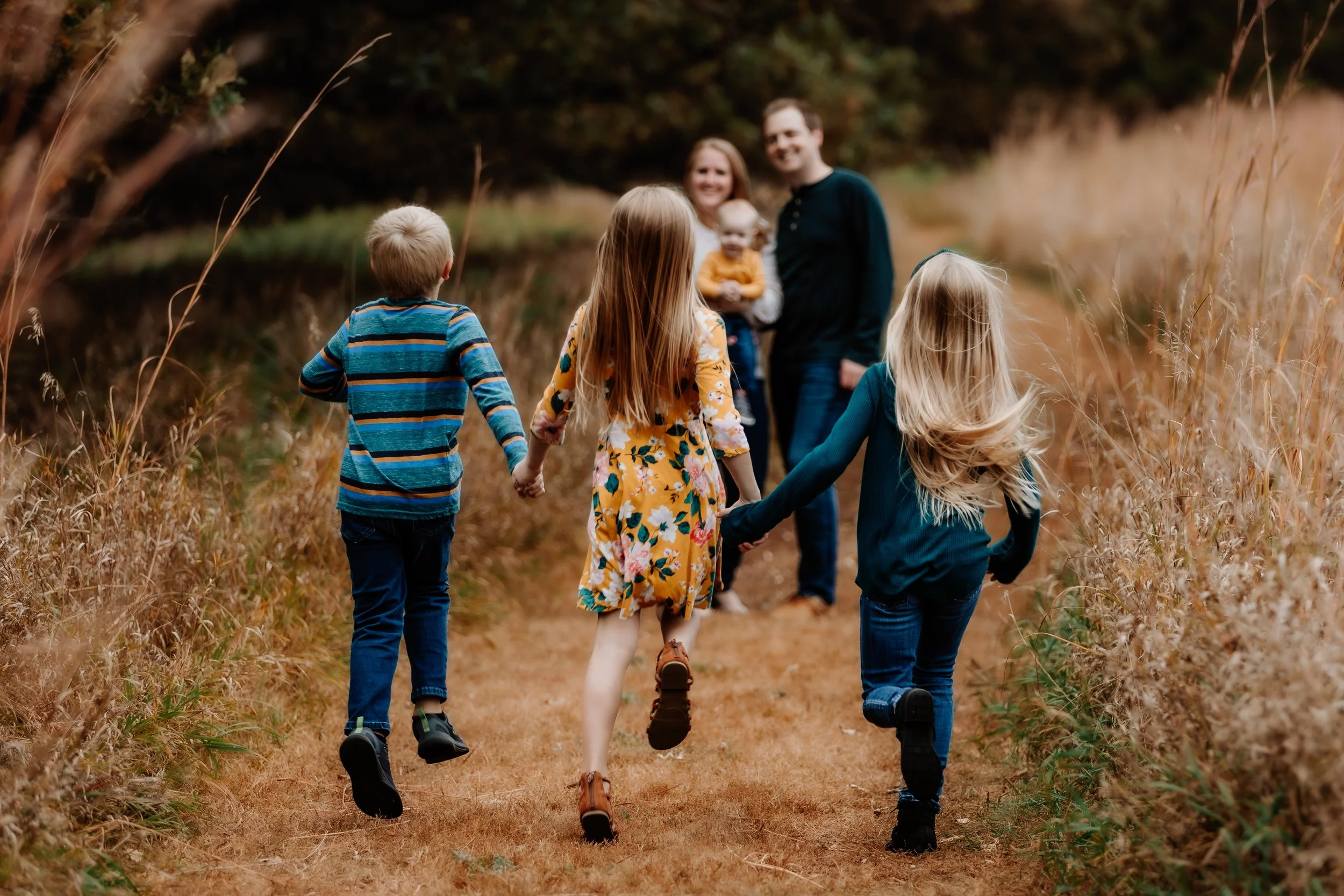 A family of six holding hands running through a grassy path in a nature setting with tall, brown grass and trees in Sioux Falls, South Dakota at Good Earth State Park.