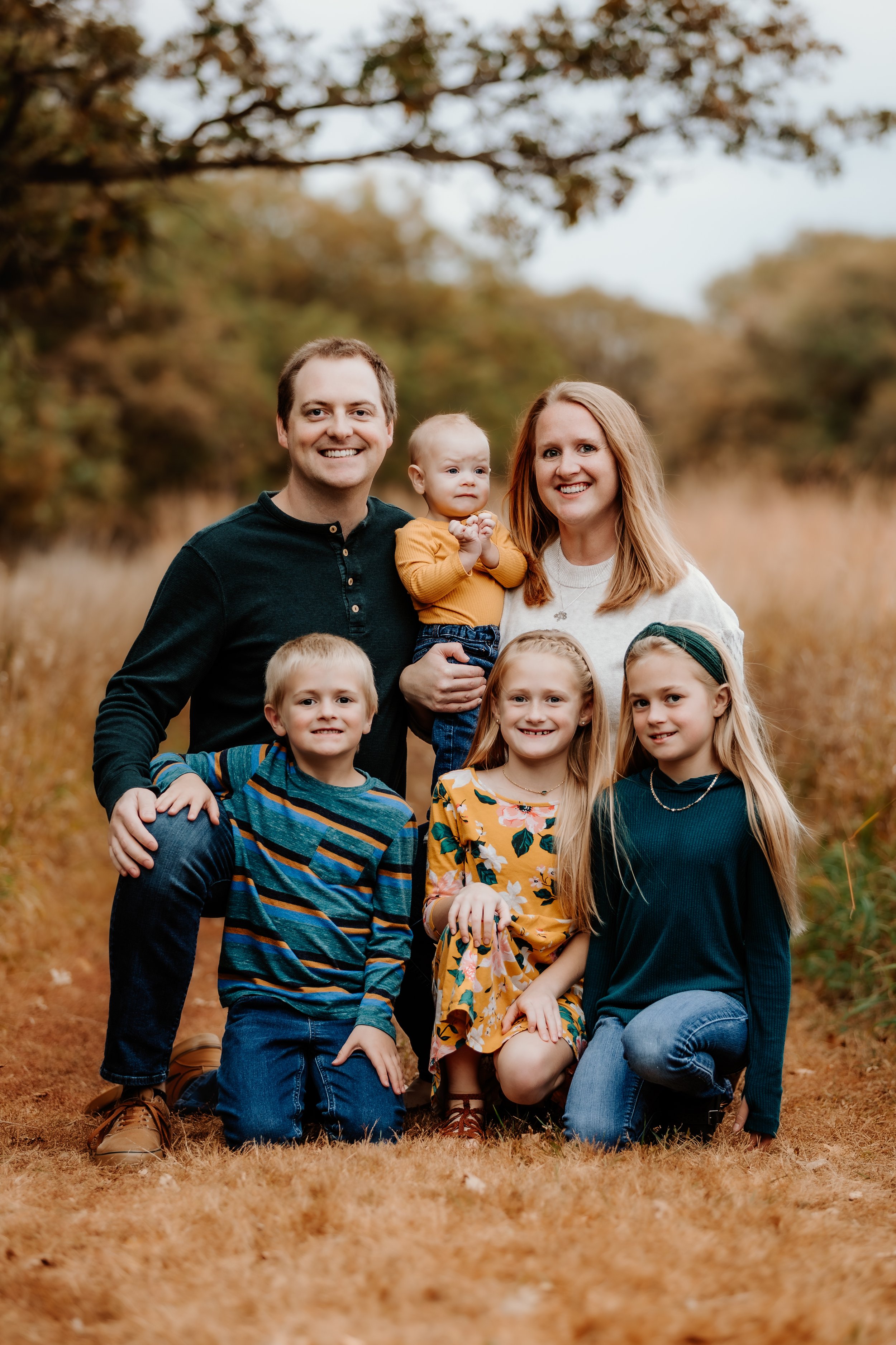 Family portrait of six people, including two adults and four children, outdoors in an autumn park with trees and grass, all smiling and posing for the photo in Sioux Falls, South Dakota at Good Earth State Park.