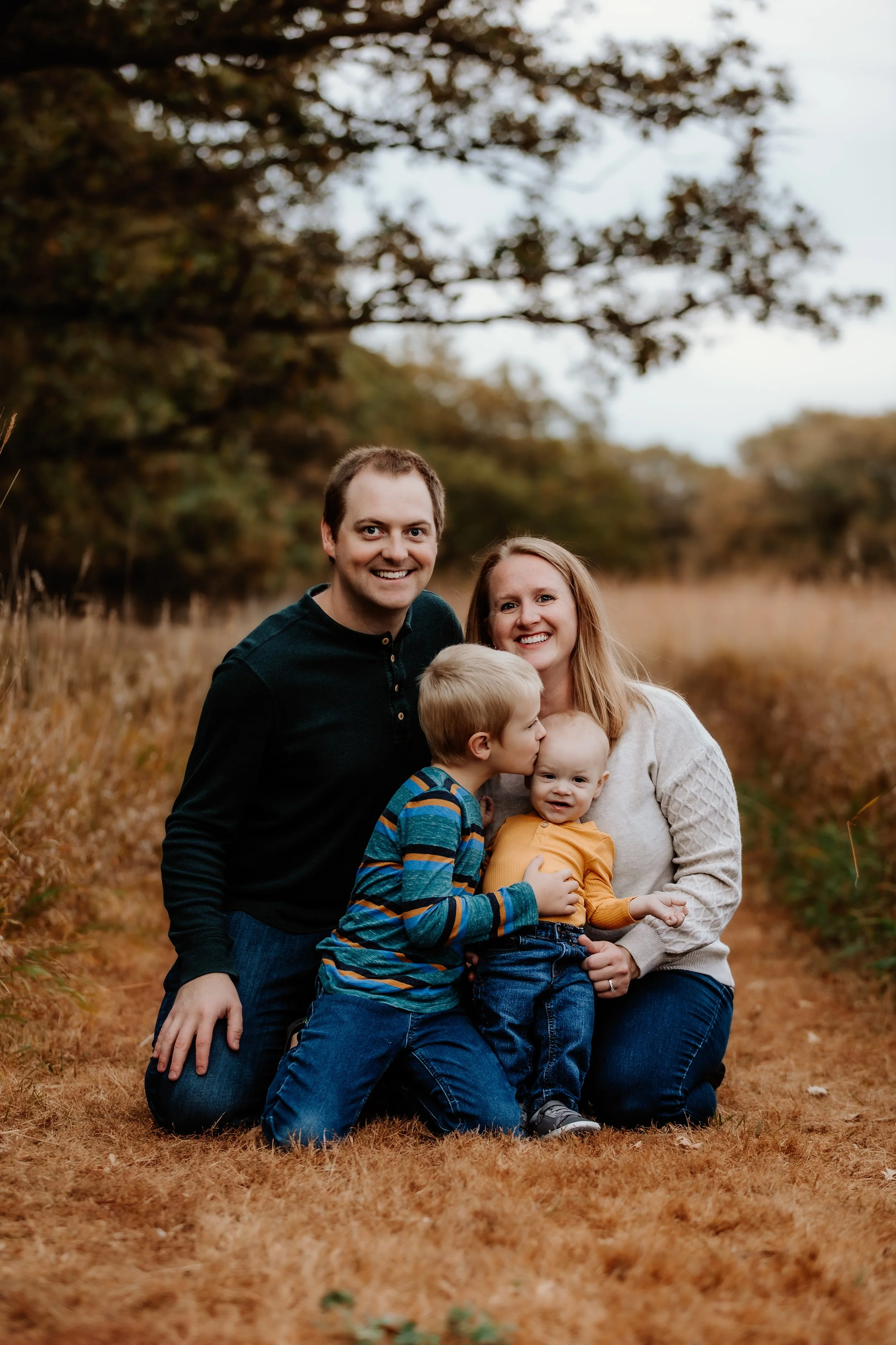 A family of four kneeling outdoors on a dirt path surrounded by trees and grass, smiling at the camera.The boy is kissing the girl on the cheek, and she is holding his arm at Good Earth State Park in Sioux Falls, South Dakota.