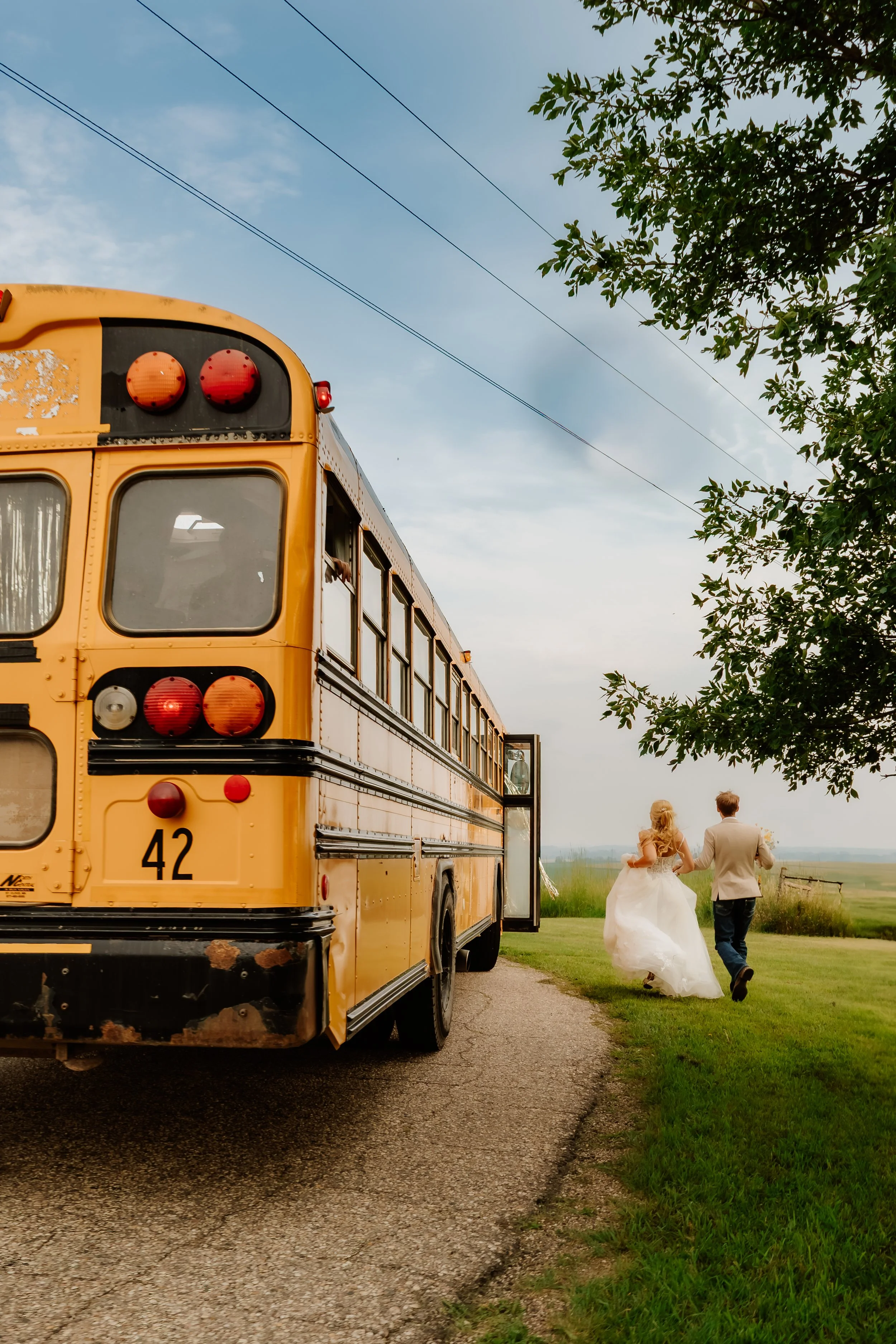 A bride and groom walking away from a yellow school bus in a rural setting, holding hands, with a tree and open fields in the background in Luverne, MN.